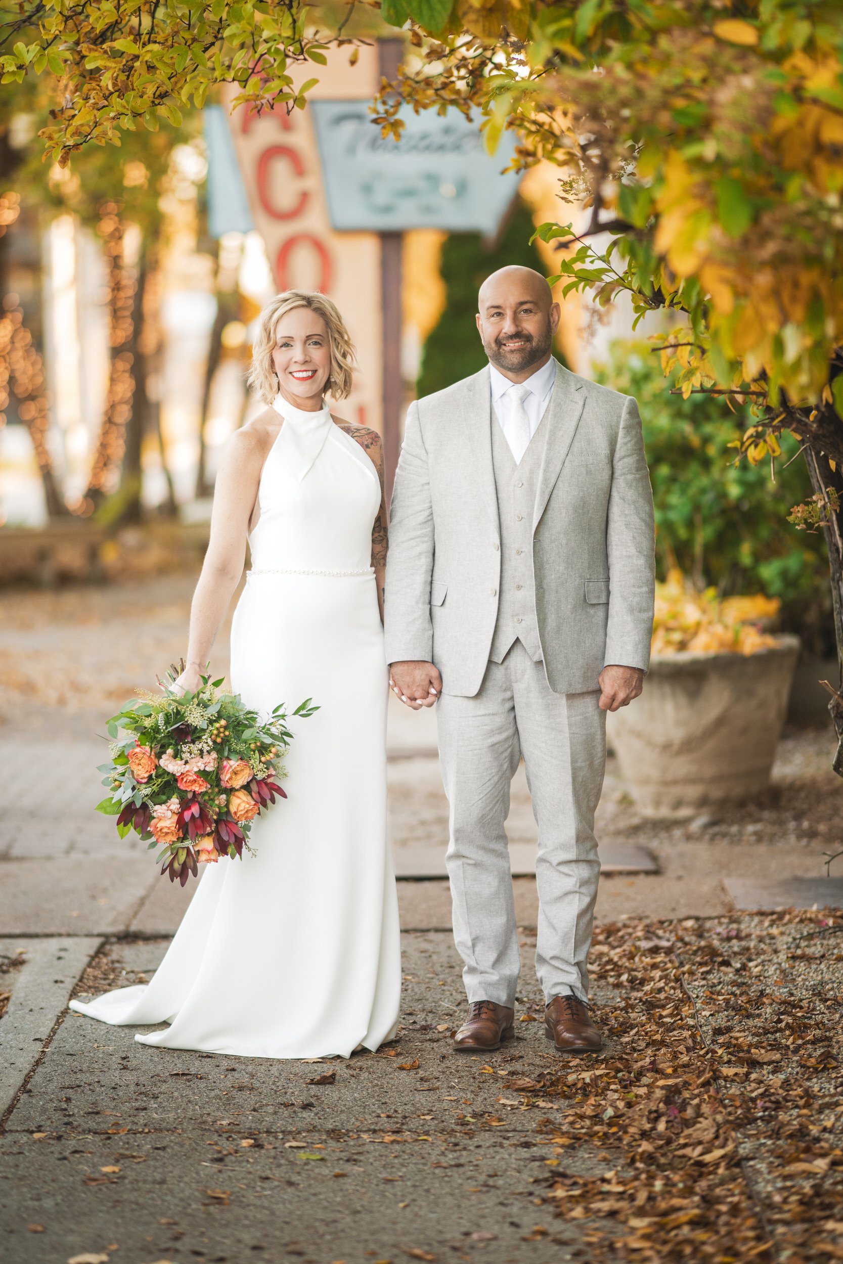 A bride in a white dress and a groom in a light gray suit stand side by side holding hands on a sidewalk lined with autumn leaves, captured by a talented Northwest Indiana and Chicago wedding photographer.