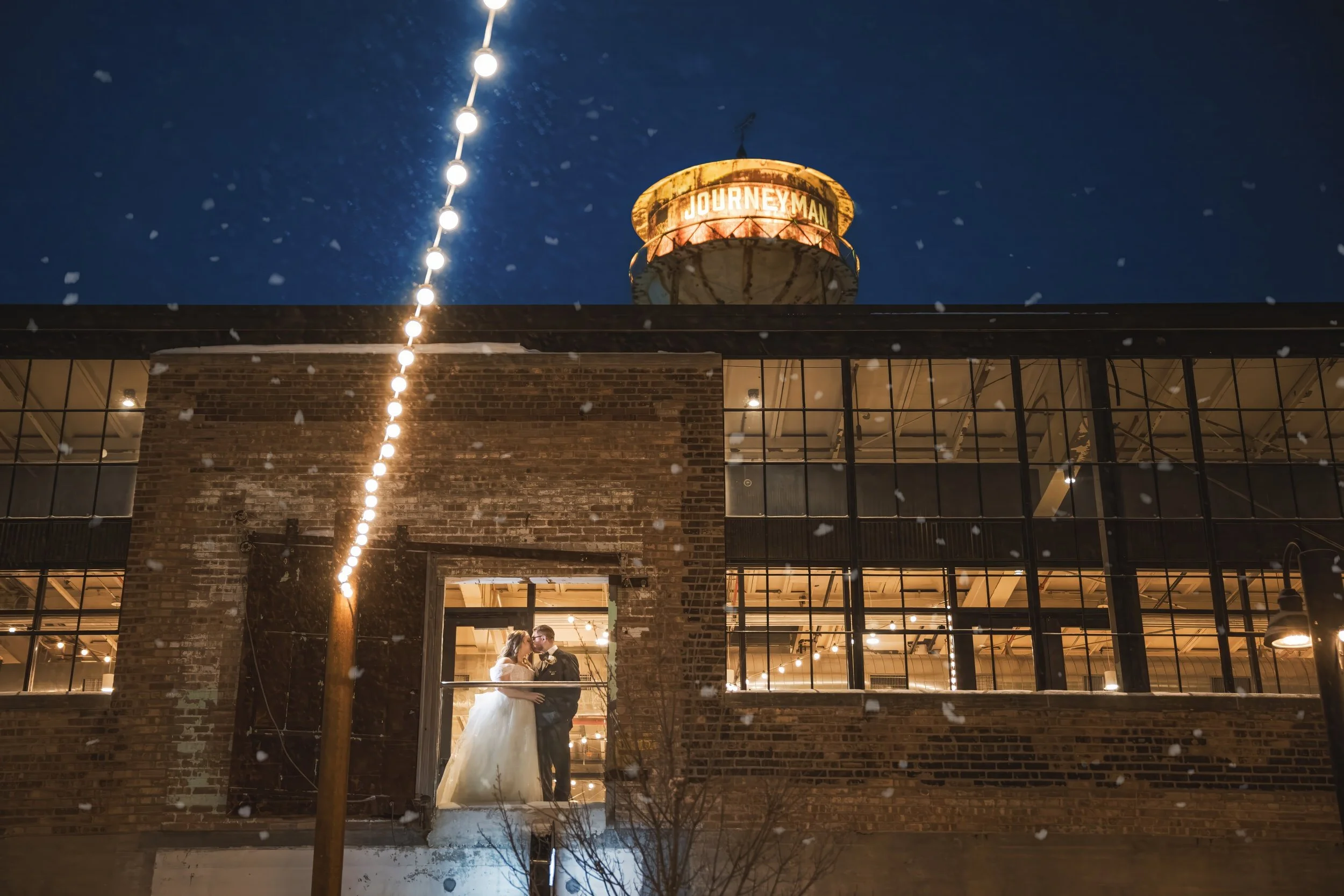 Snow falls in the night sky as the bride and groom kiss on the balcony with the Journeyman tower lit in the background.