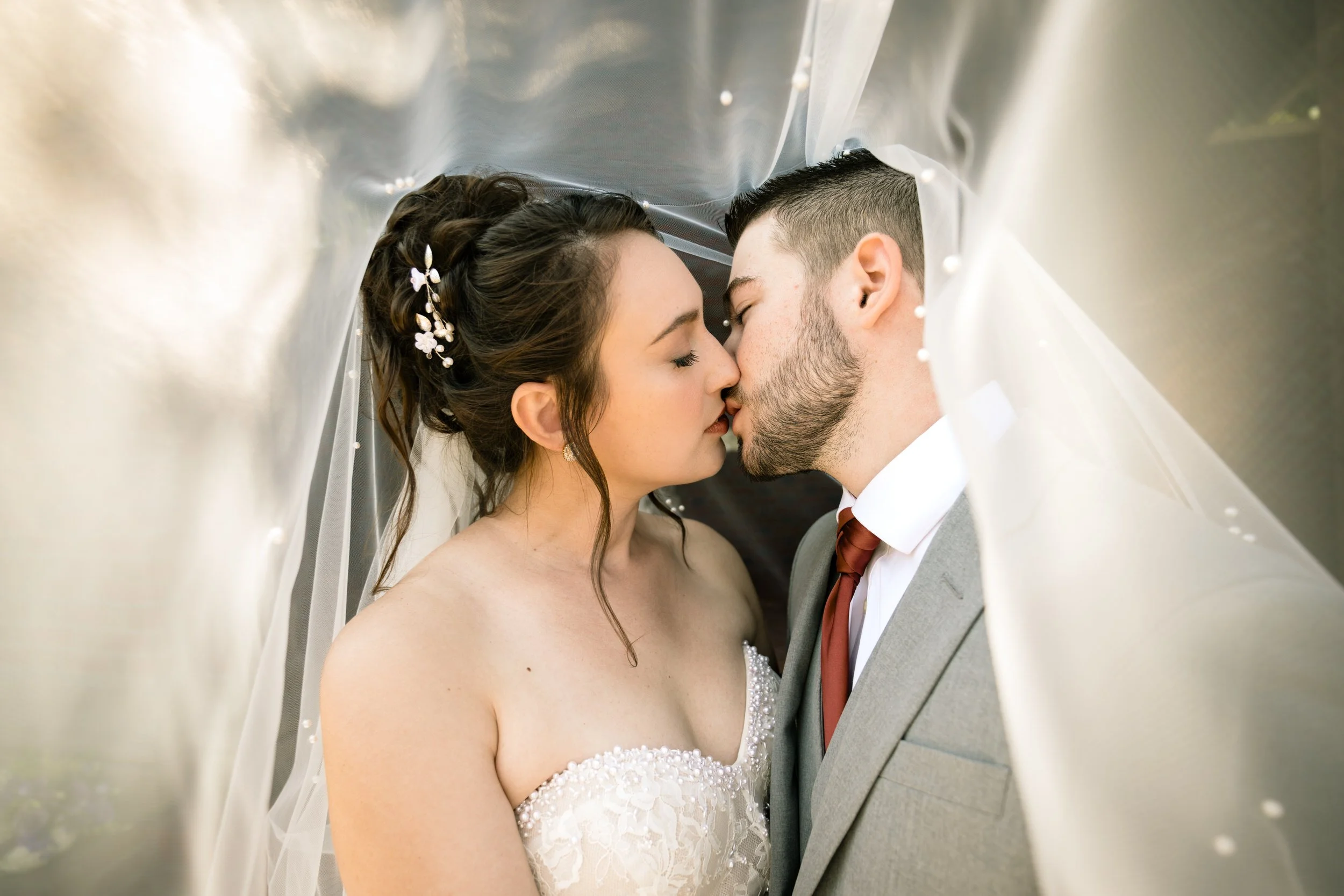 A bride and groom kiss under a sheer wedding veil, captured beautifully by a northwest Indiana and Chicago wedding photographer; the bride wears a strapless dress and the groom is in a suit with a red tie.