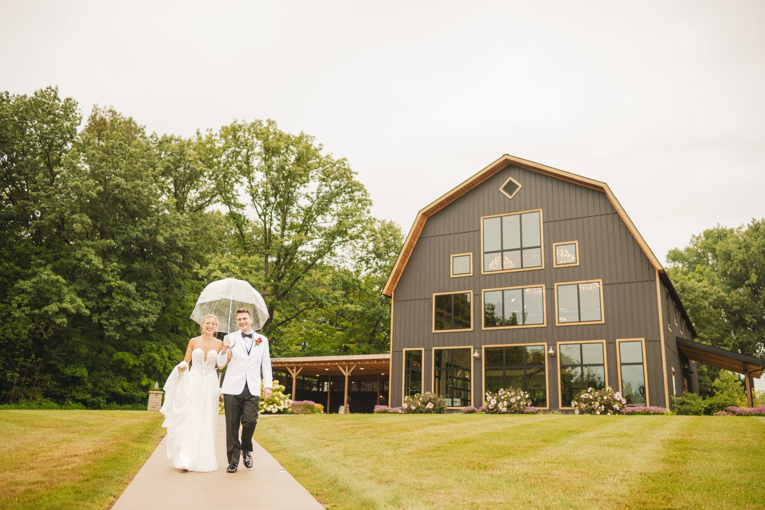 A bride and groom walk in the rain holding a clear umbrella with White Oak Farm Venue in the background.