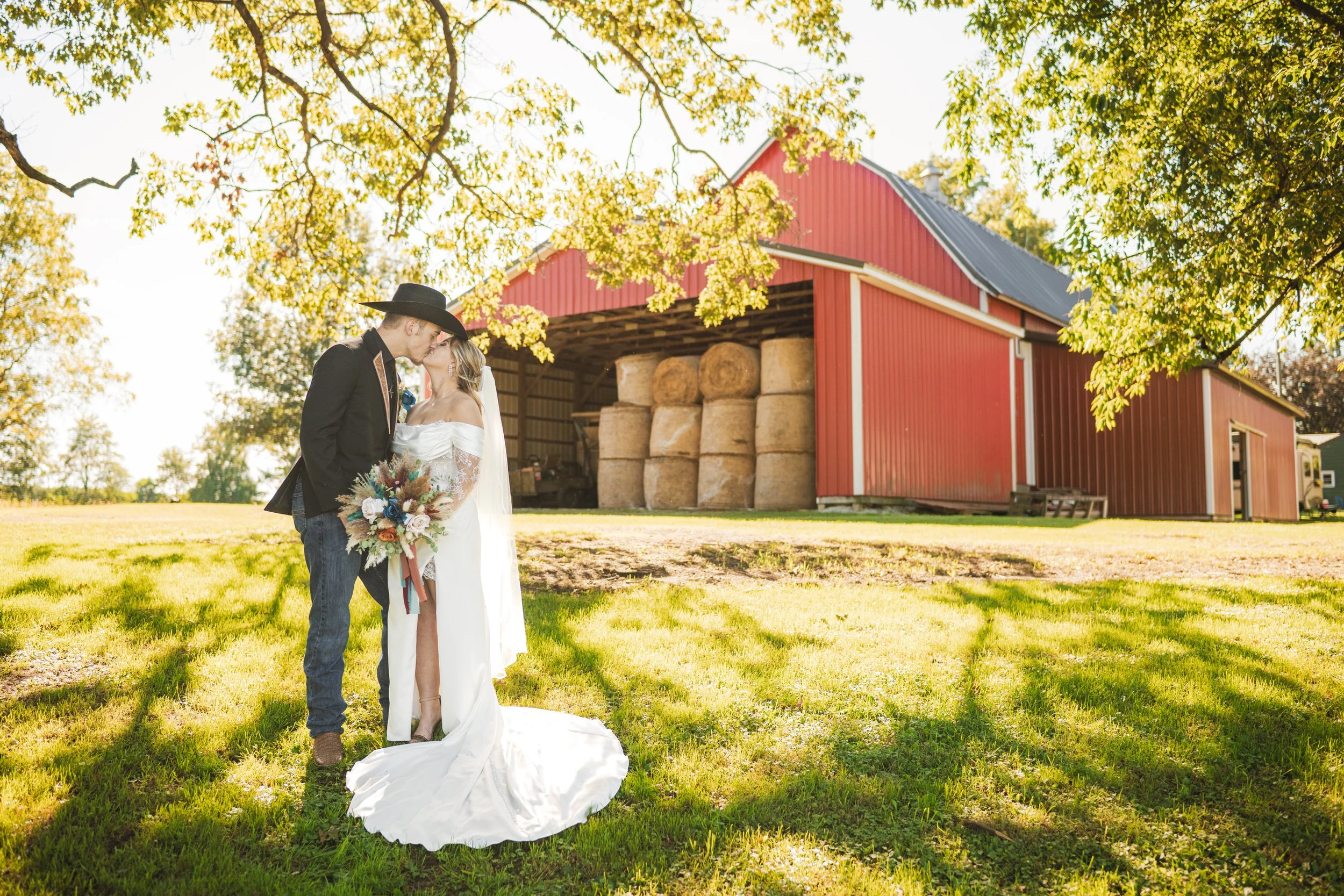 A broom in a cowboy hat kisses his bride with a red barn behind them.