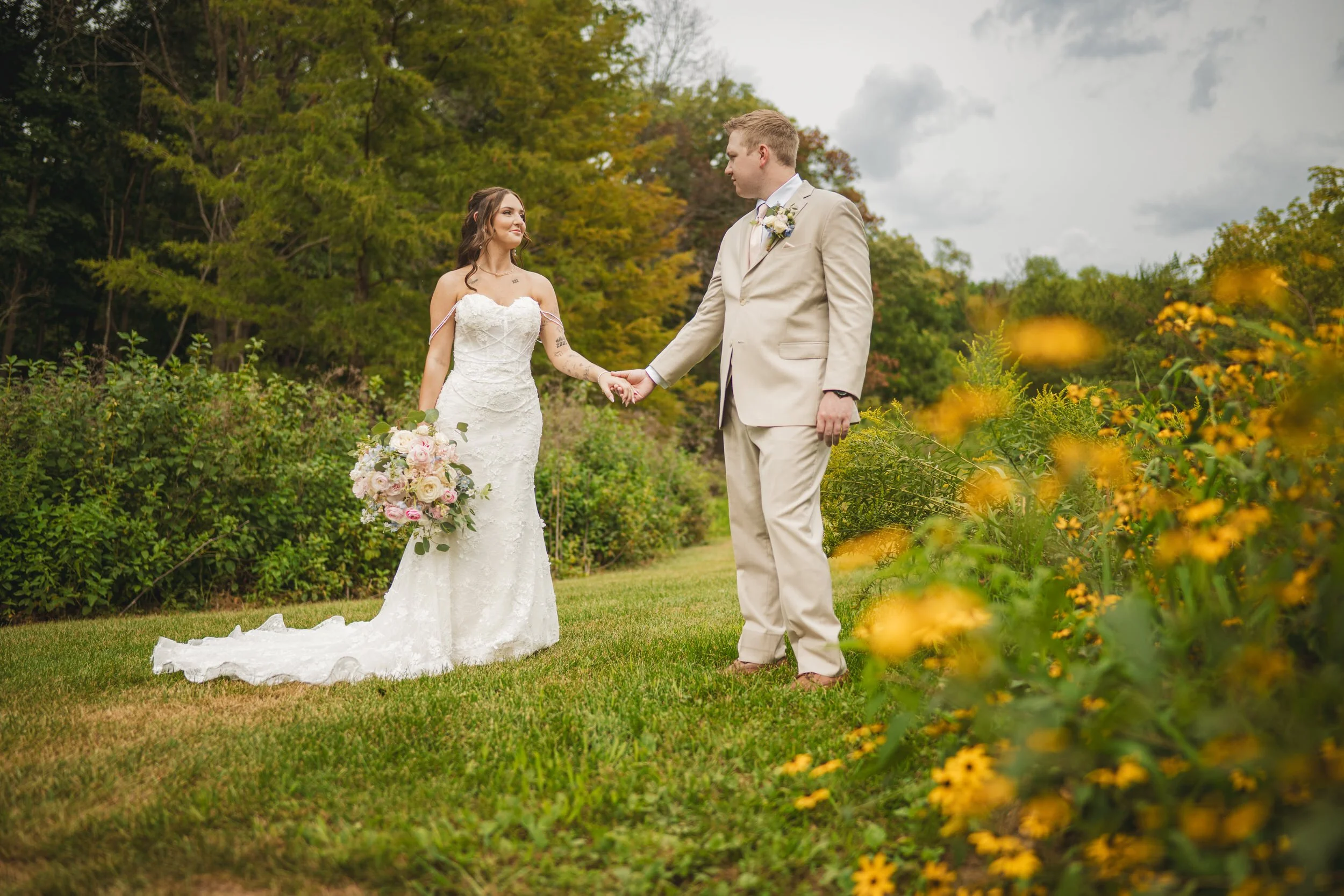 Yellow flowers are in the foreground as the bride and groom gaze at each other with puffs of clouds in the sky above them.