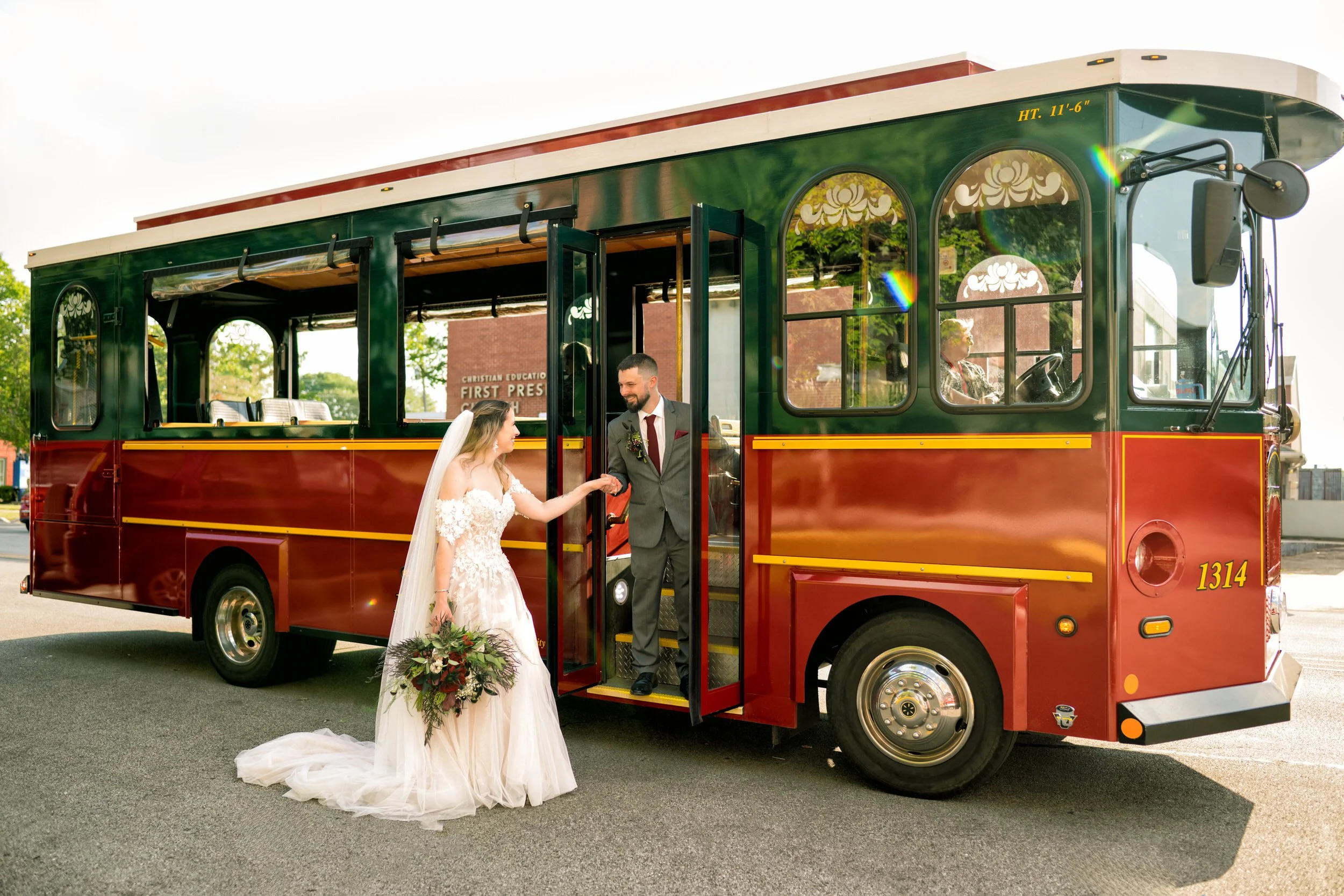 A bride in a wedding dress holds a bouquet and steps off a red trolley bus with the assistance of her groom, captured by a northwest Indiana and Chicago wedding photographer.