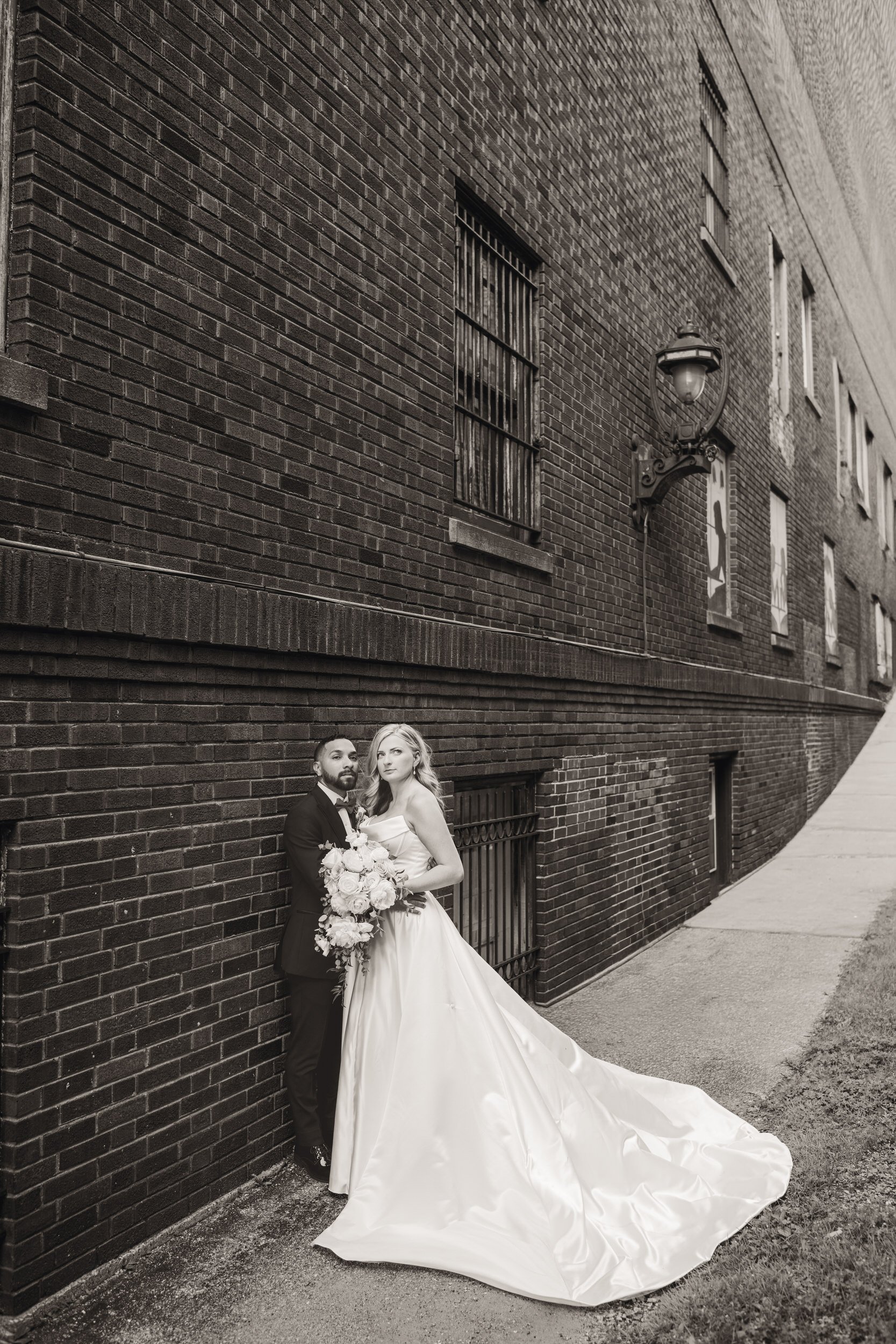 A cement path leads toward the bride groom as they lean up against a brick wall after their wedding in the Crown Point Square.