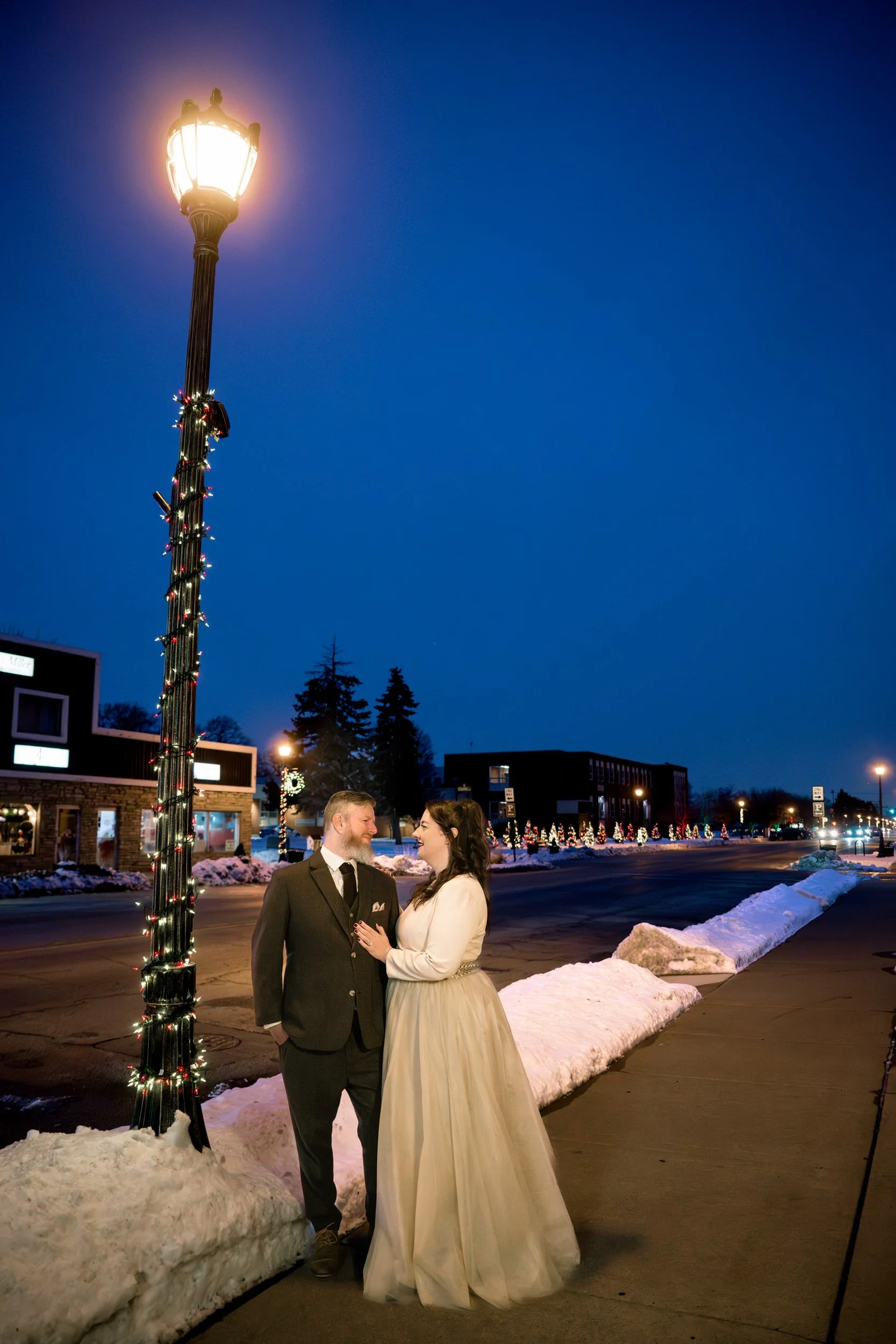 A couple dressed formally stands on a snowy sidewalk at dusk beside a lit streetlamp, with buildings and holiday lights in the background, captured by a Northwest Indiana and Chicago wedding photographer.