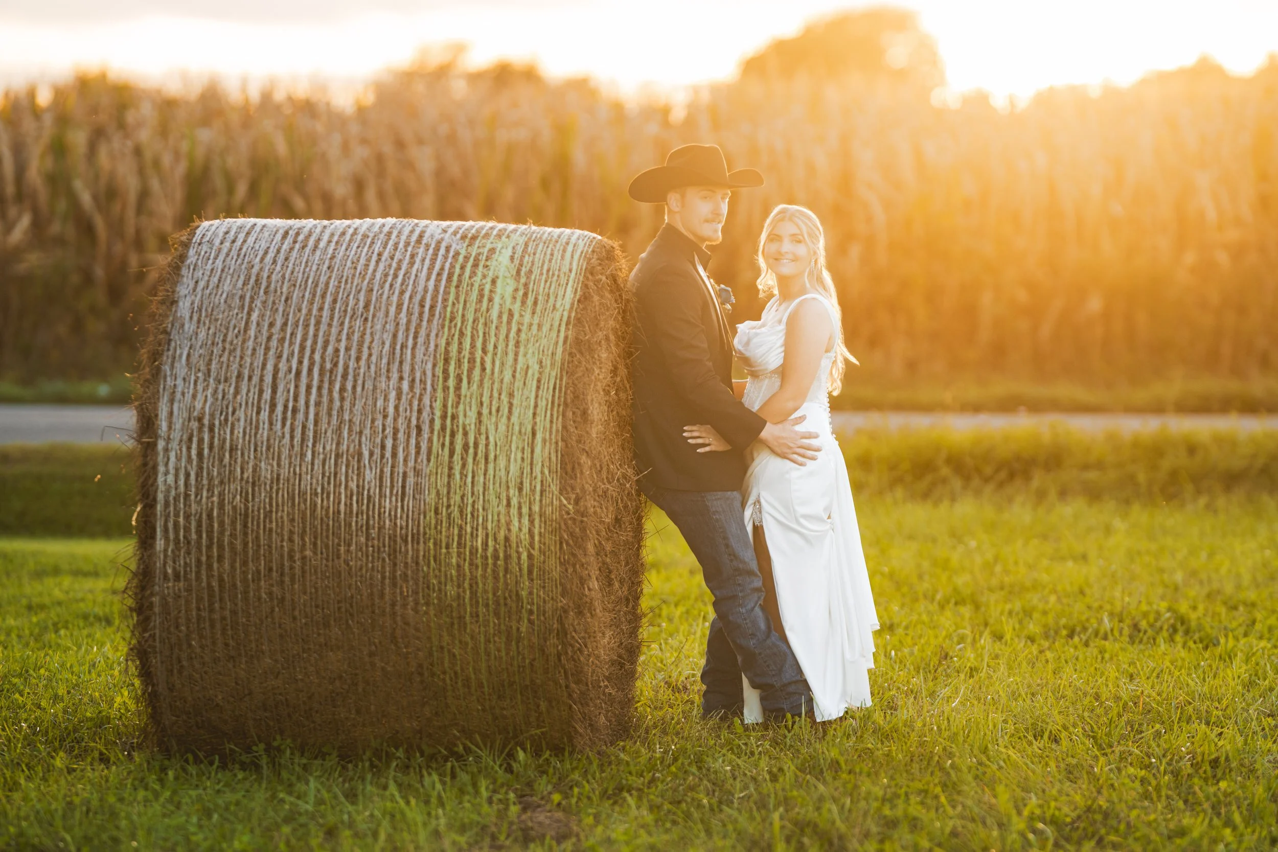 A bride and groom lean against a hay bale as the sun sets behind a cornfield.
