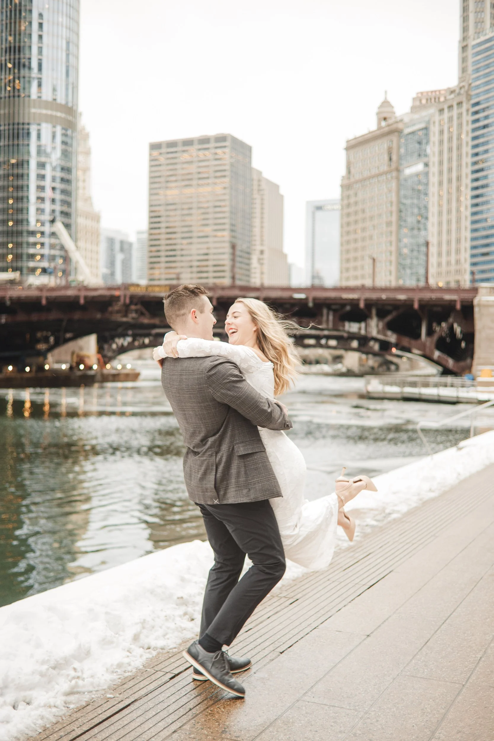 A groom to be spins his fiance around on the Chicago Riverwalk with skyscrapers behind them.
