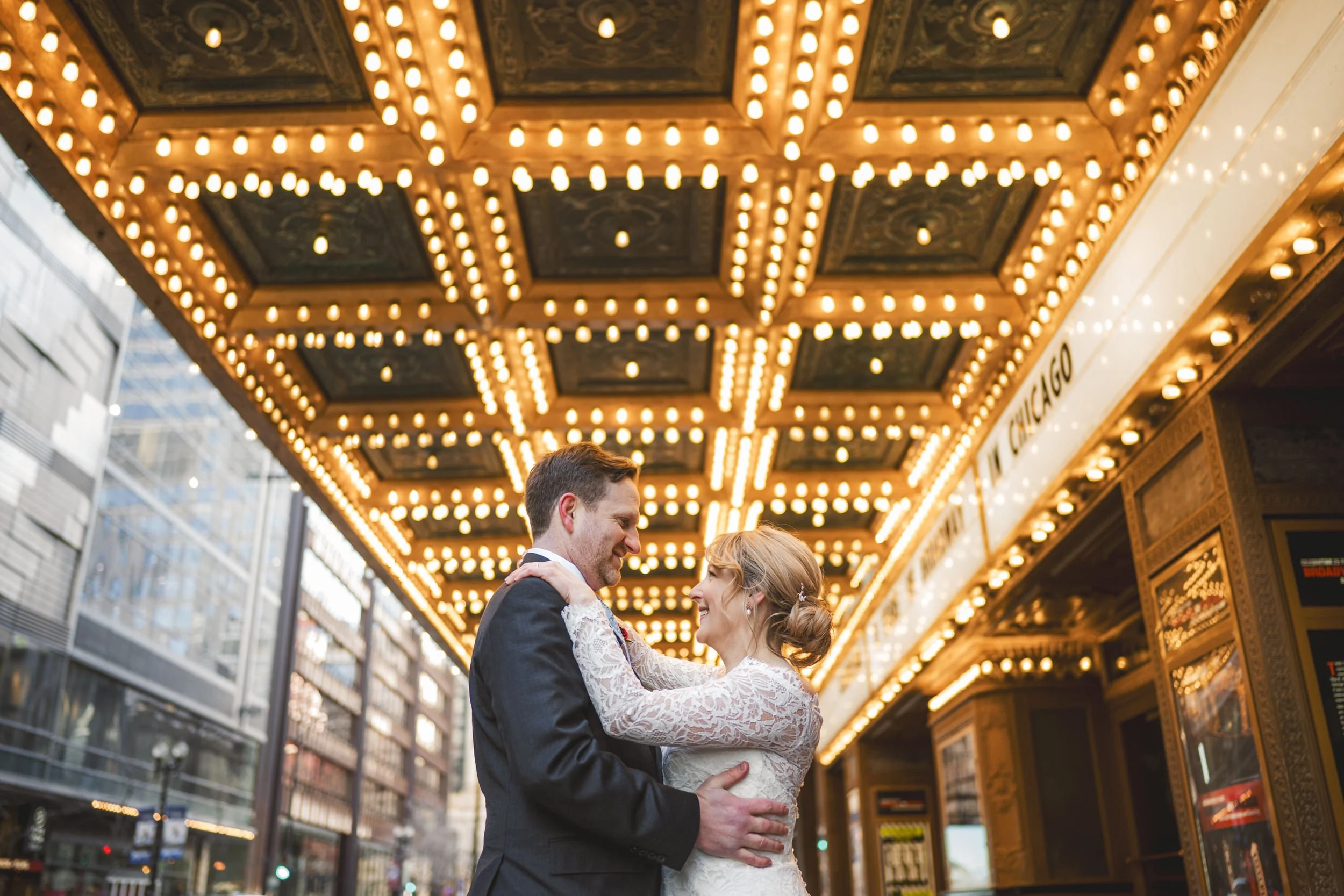 A bride and groom embrace and smile at each other beneath a brightly lit theater marquee in an urban setting, captured by a Northwest Indiana and Chicago wedding photographer.