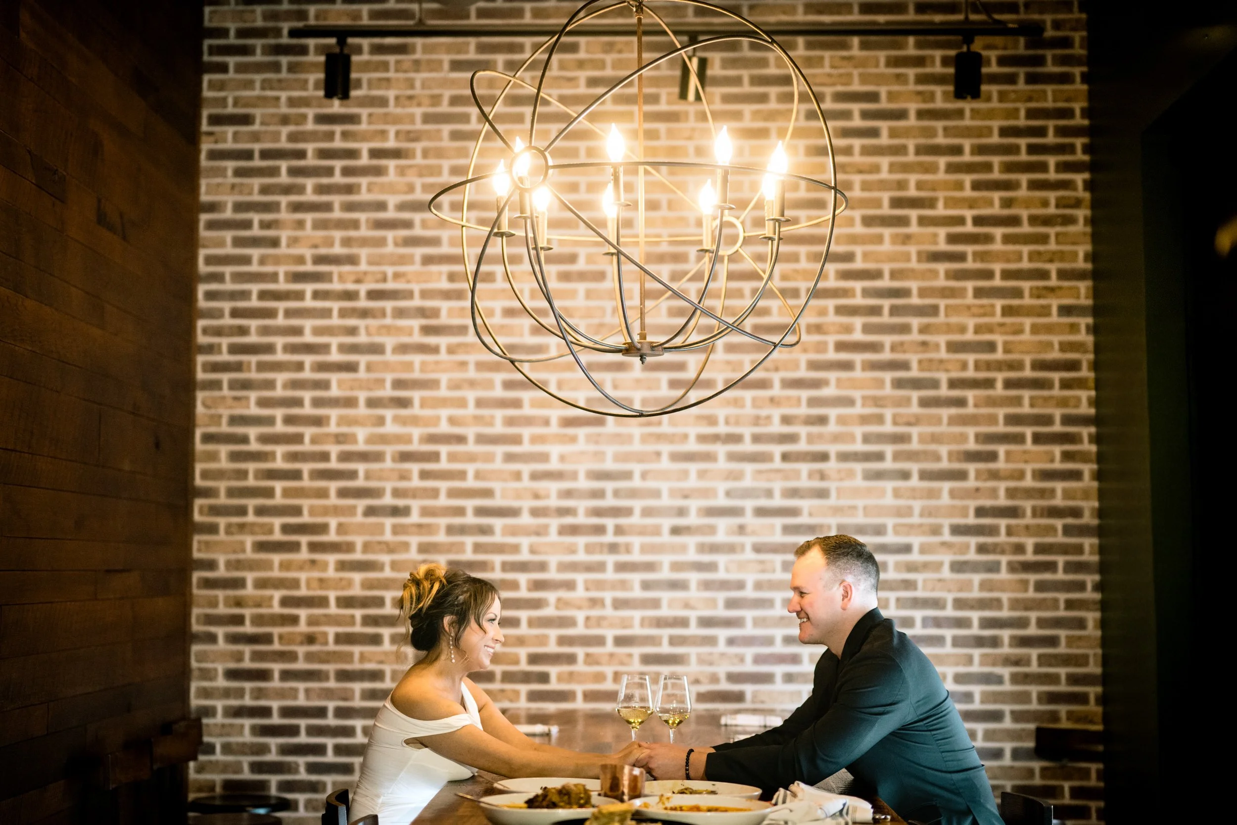 An engaged couple have dinner underneath a golden chandelier at a high end restaurant.