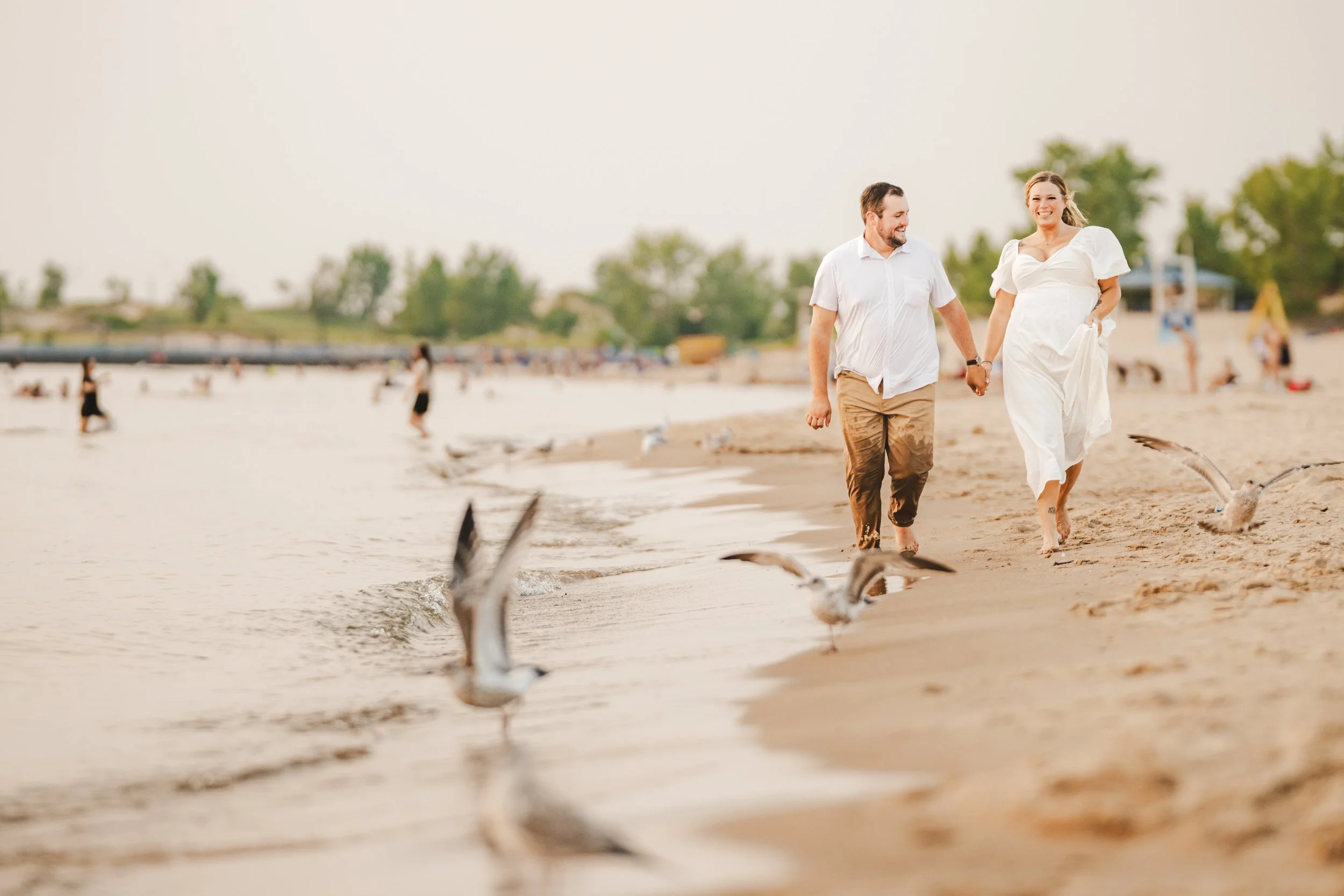 A man and woman holding hands walk barefoot along Silver Lake Beach at golden hour, with seagulls nearby and people in the background near the water—a timeless scene beautifully captured by a skilled southwest Michigan wedding photographer.