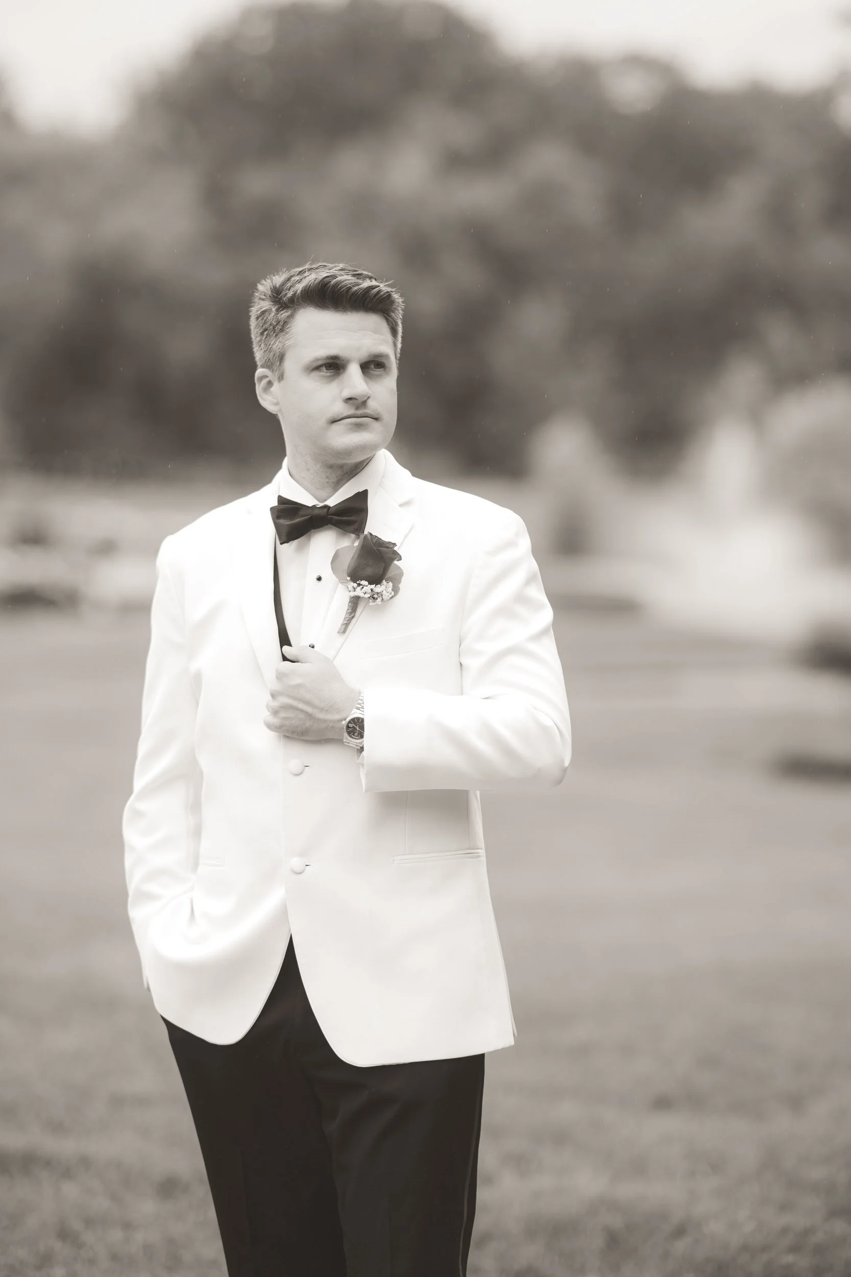 A black and white image of a groom holding his lapel and looking off into the distance as light rain falls.