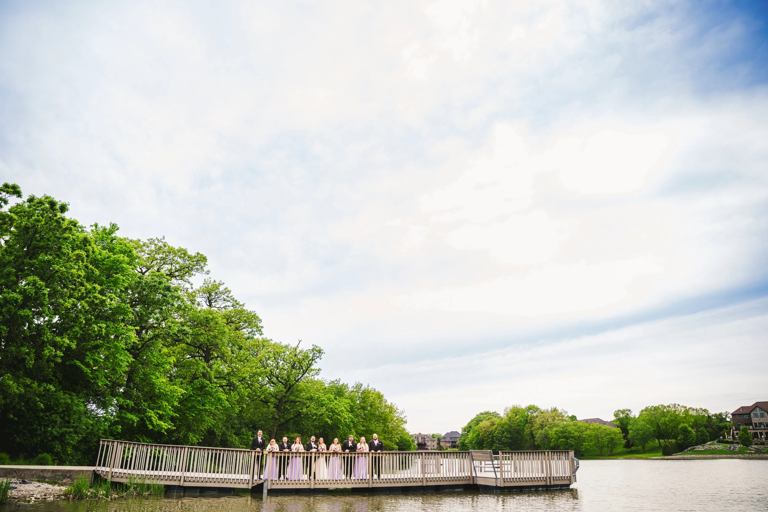 A wedding party stands on the pier overlooking the lake at Lake Hills Park in St. John, Indiana with a blue sky above them.