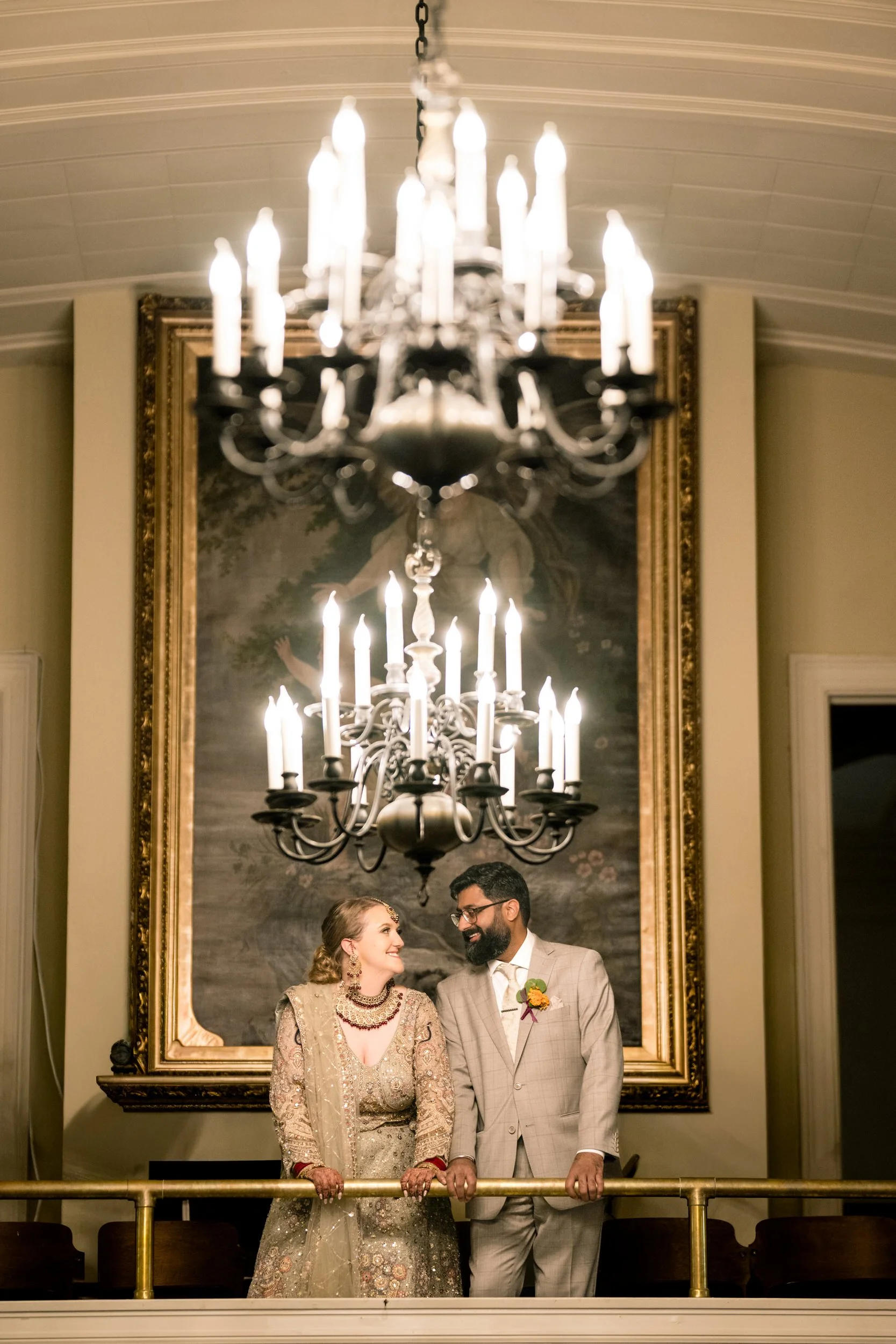 A couple dressed in formal attire stands together, smiling at each other beneath a lit chandelier, with a large framed painting in the background—captured by a skilled northwest Indiana and Chicago wedding photographer.