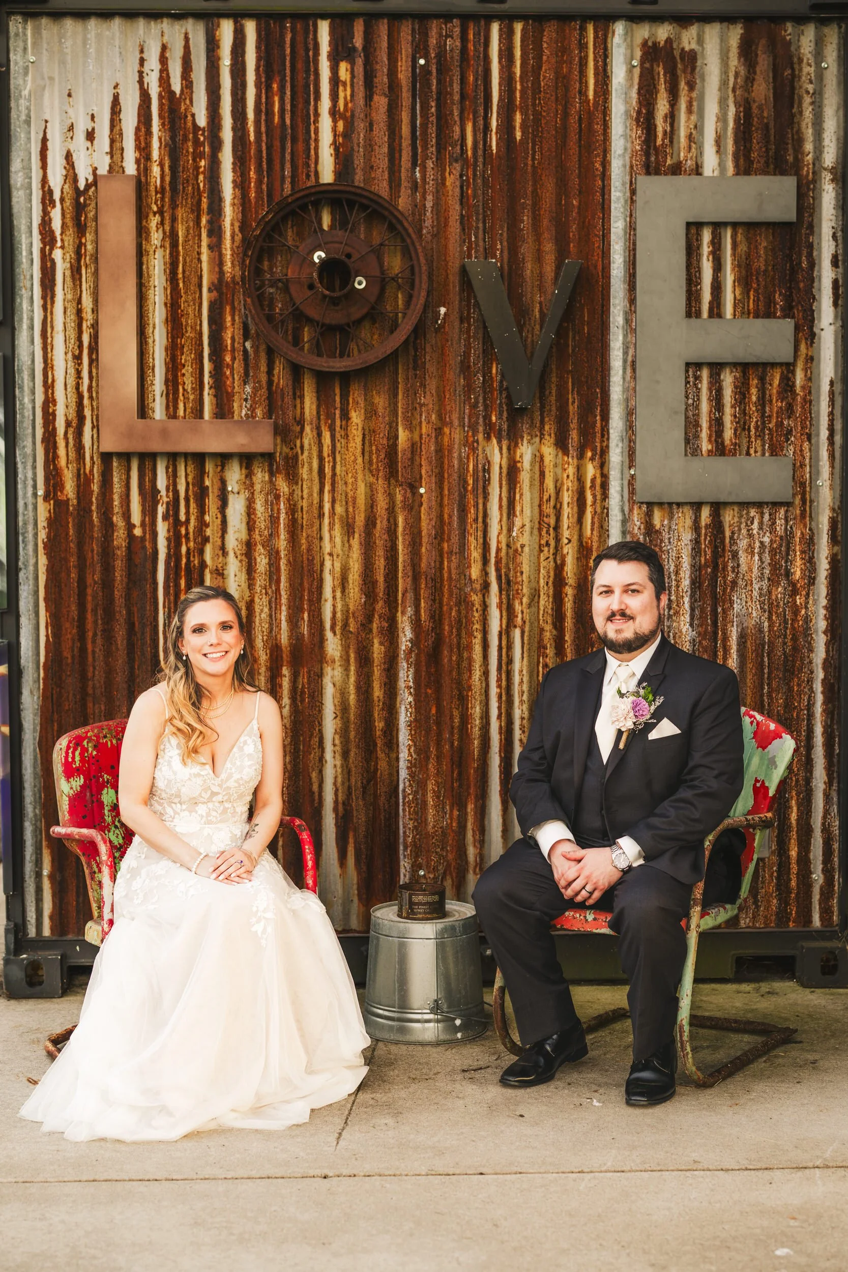 A rustic metal sign that spells out L-O-V-E is shown behind a bride and groom at their rustic wedding venue The Design Barn.