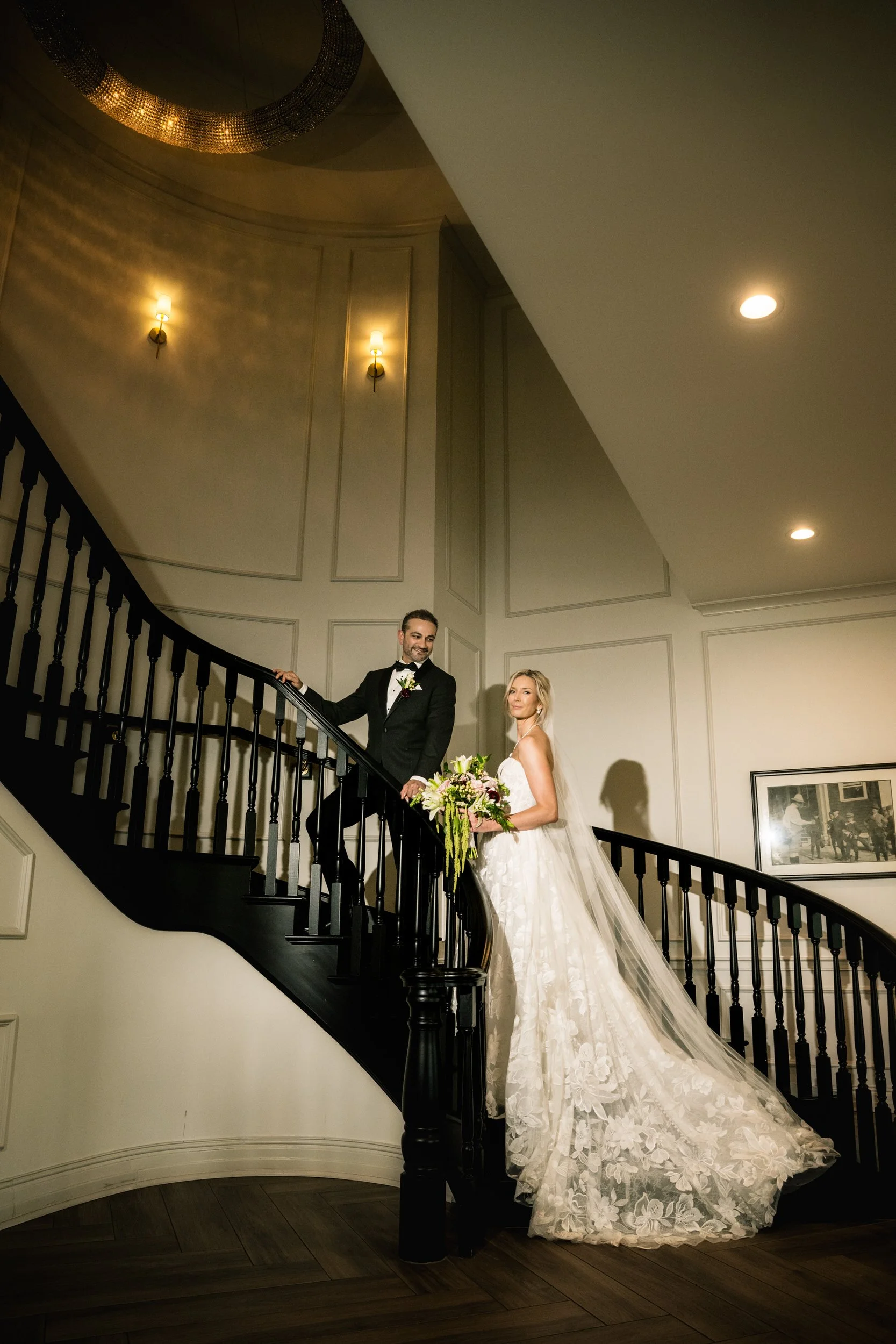 The bride's train flows down the staircase at Midlothian Country Club as she looks at the camera and groom looks at her.