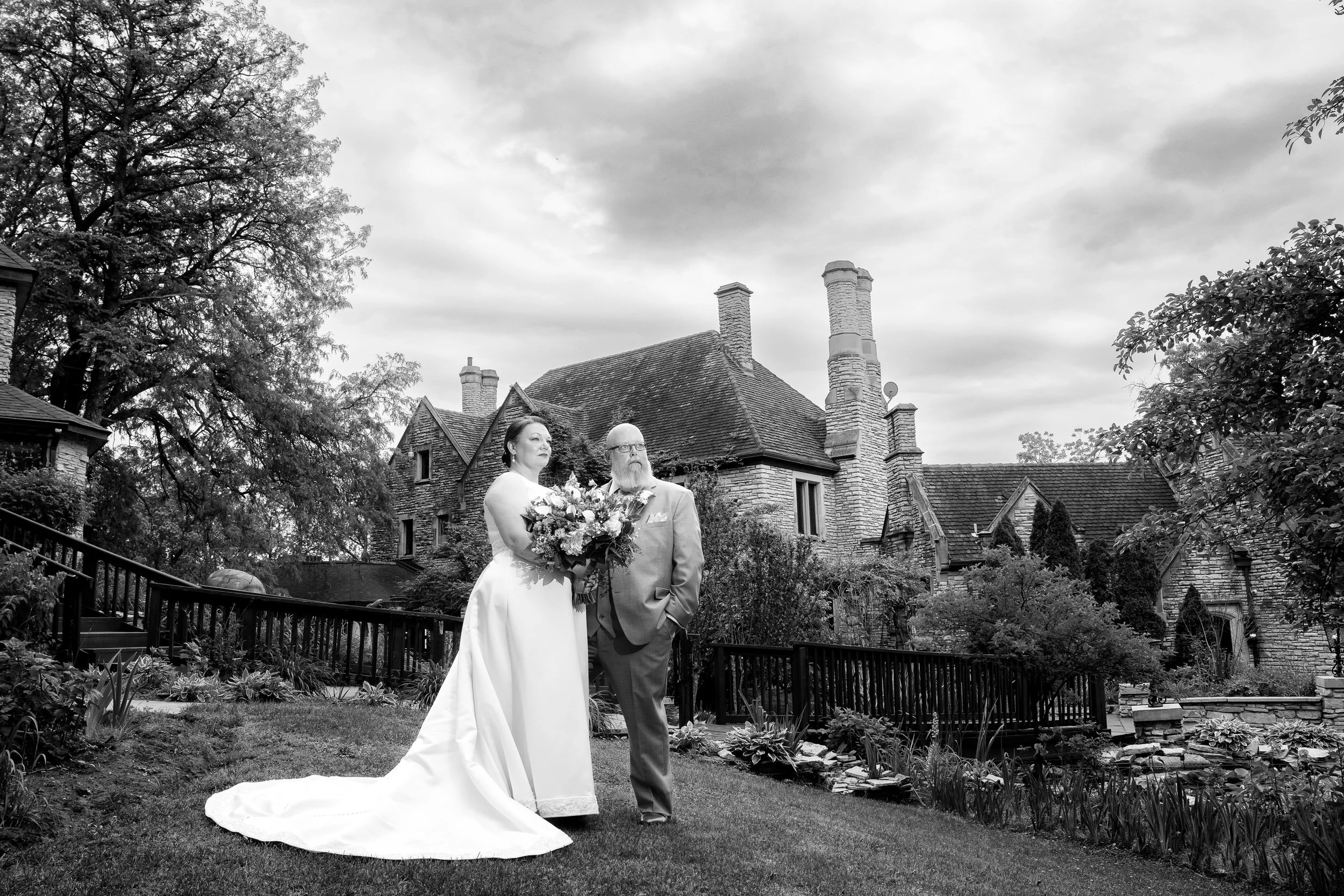 A bride in a white gown holding a bouquet walks beside an older man in a suit, outside a large stone house on a cloudy day, beautifully captured by a northwest Indiana and Chicago wedding photographer.