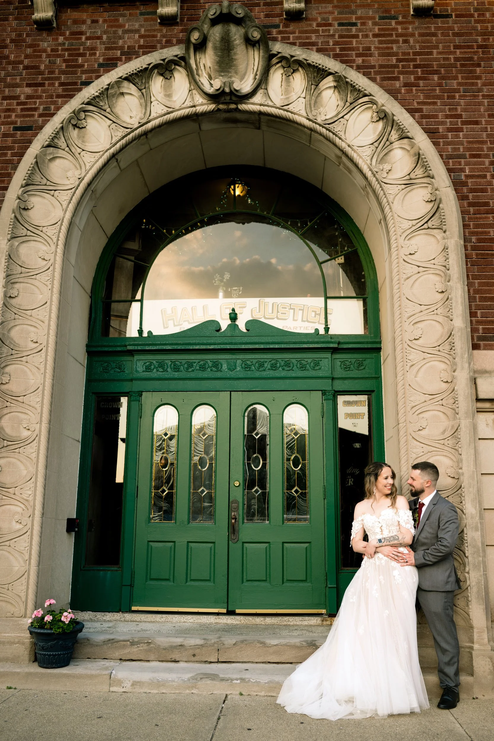 A couple in wedding attire stands before a green door under the "Hall of Justice" arch on a brick building, captured by a Northwest Indiana and Chicago wedding photographer.