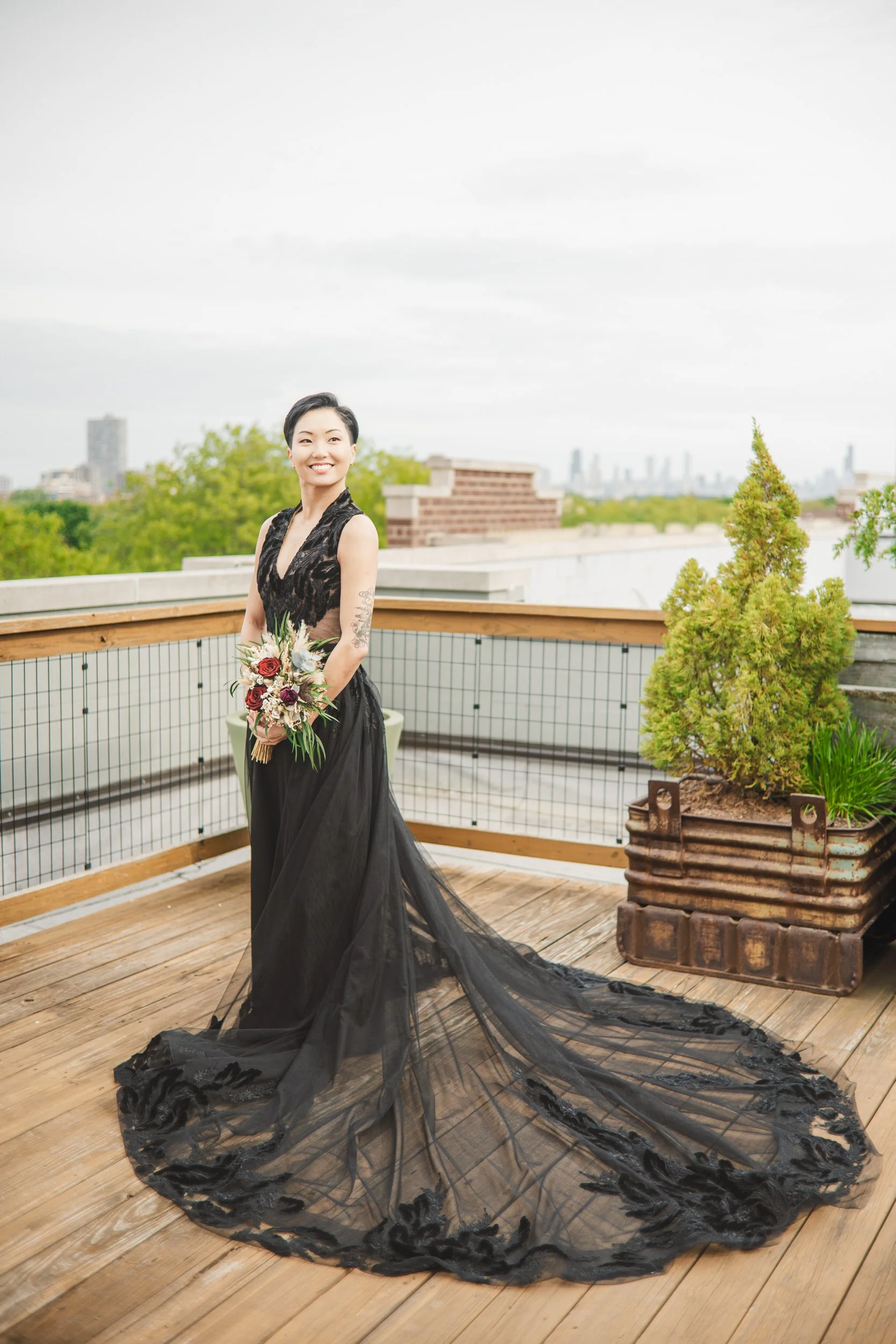 A bride stand on a Chicago rooftop in a black wedding dress, smiling and holding flowers with the city of Chicago in the background.