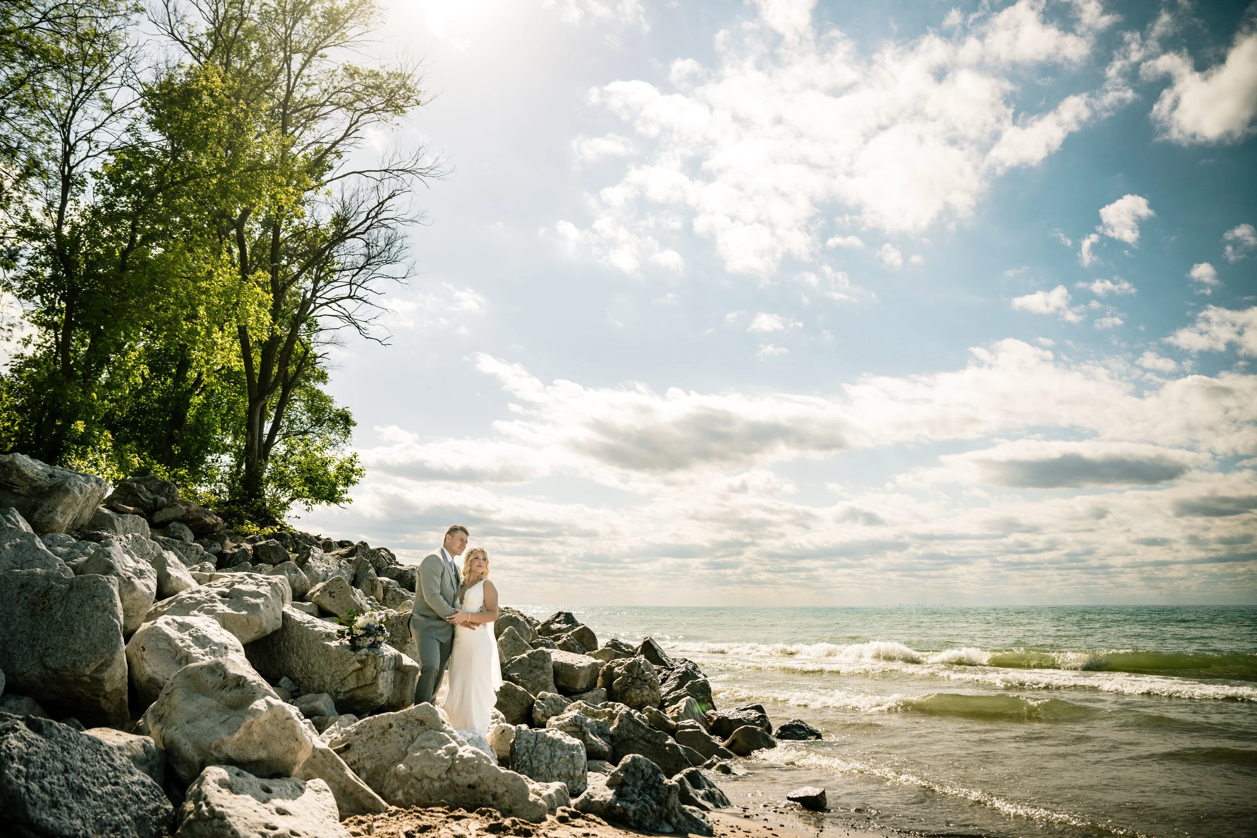 A couple in wedding attire stands on a rocky shoreline near trees, with waves and a partly cloudy sky in the background—captured beautifully by a Northwest Indiana and Chicago wedding photographer.