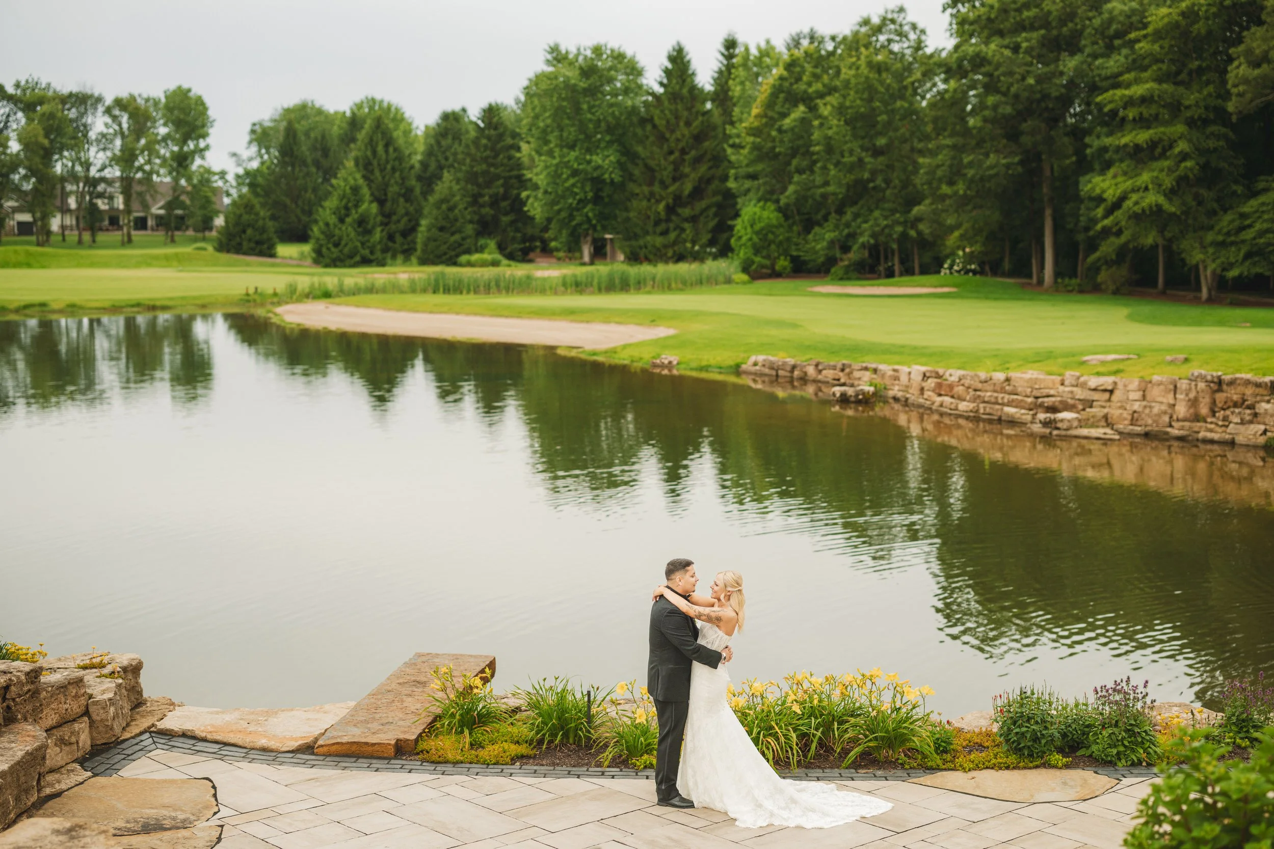 A bride and groom dance on a stone platform with a picturesque lake and Sandy Pines golf course behind them.