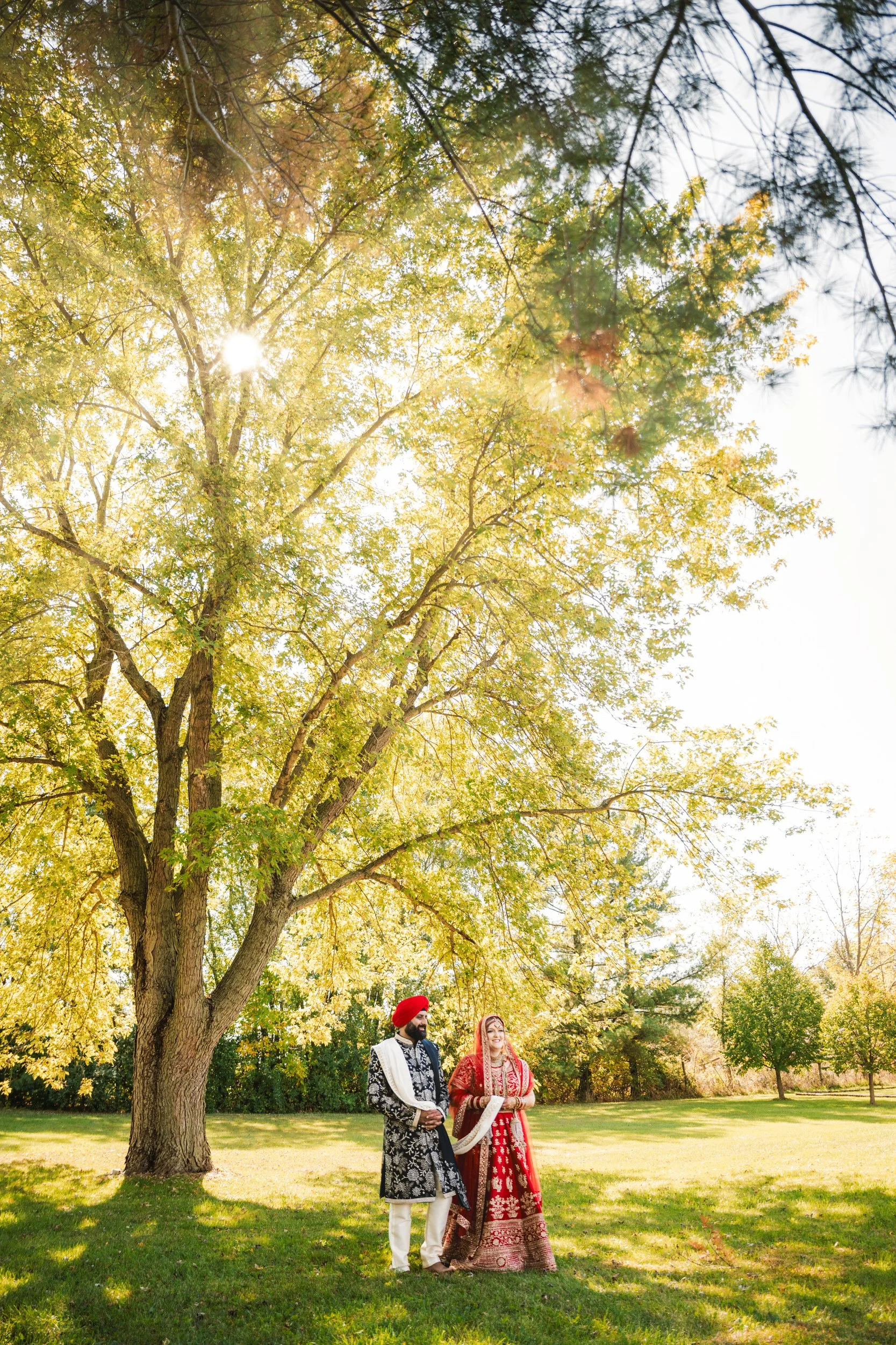 A bride and groom stand in radiant Indian wedding attire as sun shines upon them through tall trees.
