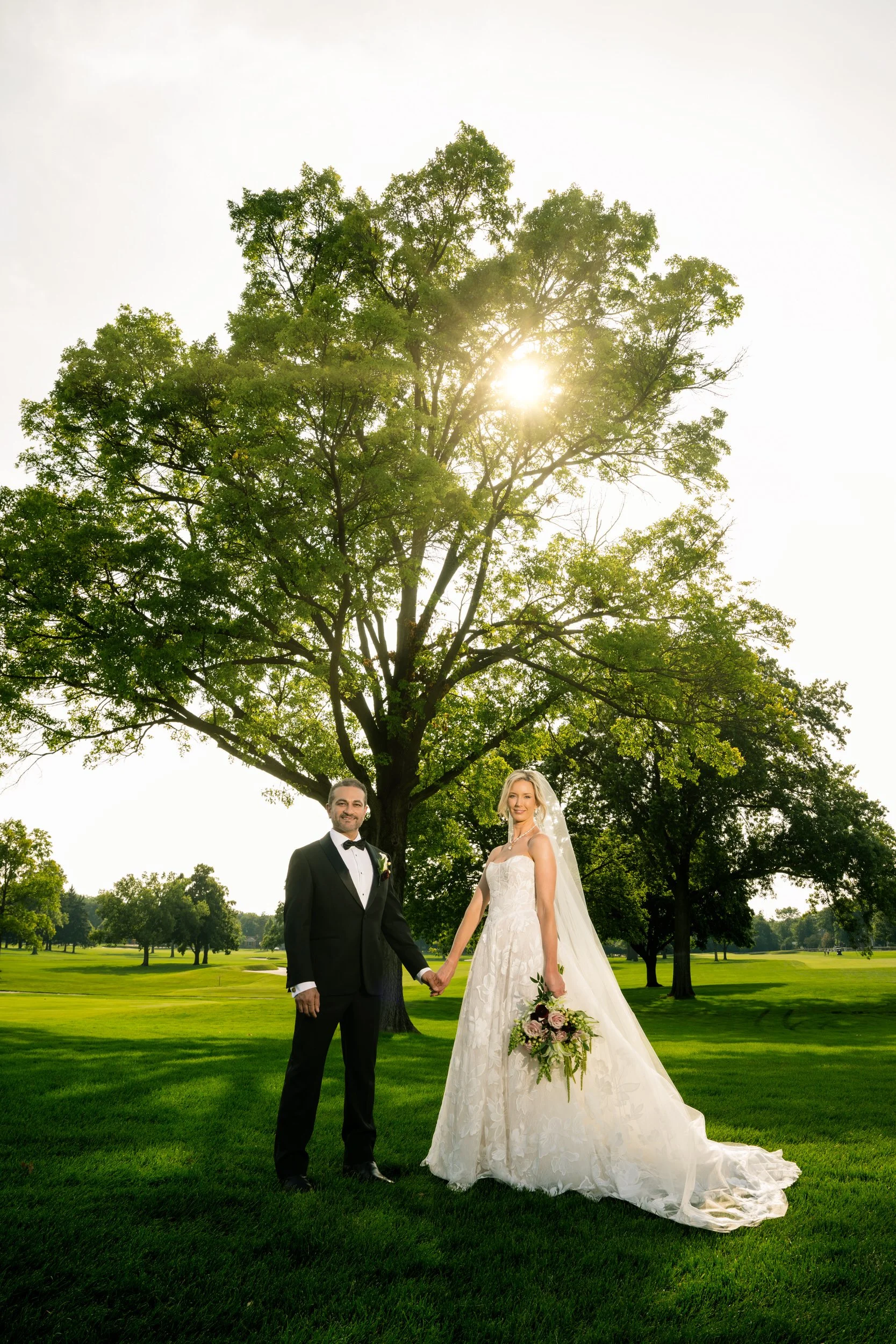 Sun bursts through the trees at Midlothian Country Club in Illinois, as the bride and groom hold hands in the shade.
