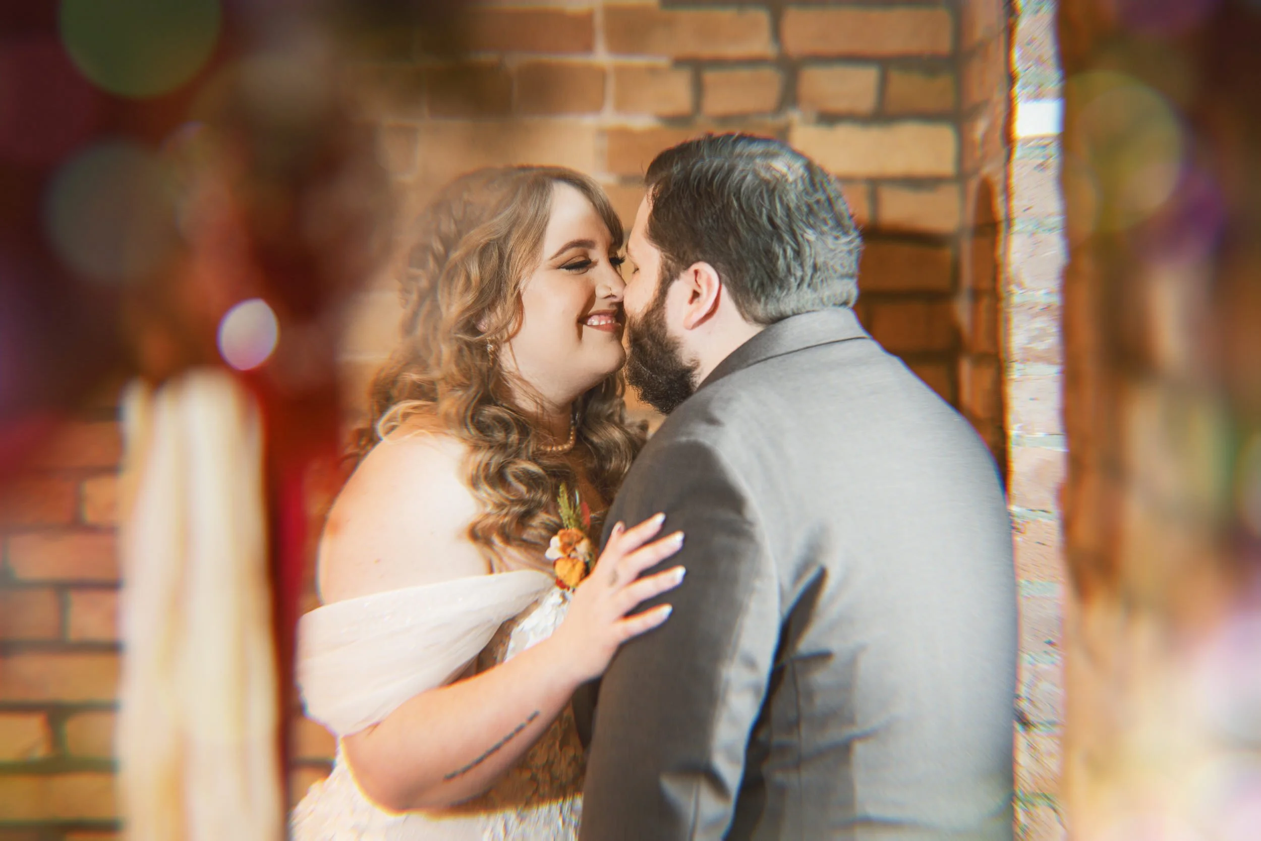 A bride and groom stand close together, smiling and about to kiss, in front of a brick wall—captured beautifully by a northwest Indiana and Chicago wedding photographer.