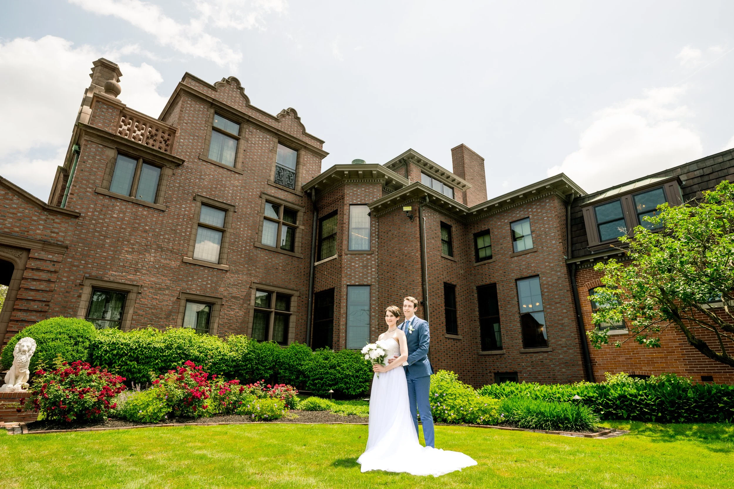 A bride and groom pose together on the lawn in front of a large brick building with landscaped gardens and greenery, captured by a Northwest Indiana and Chicago wedding photographer.