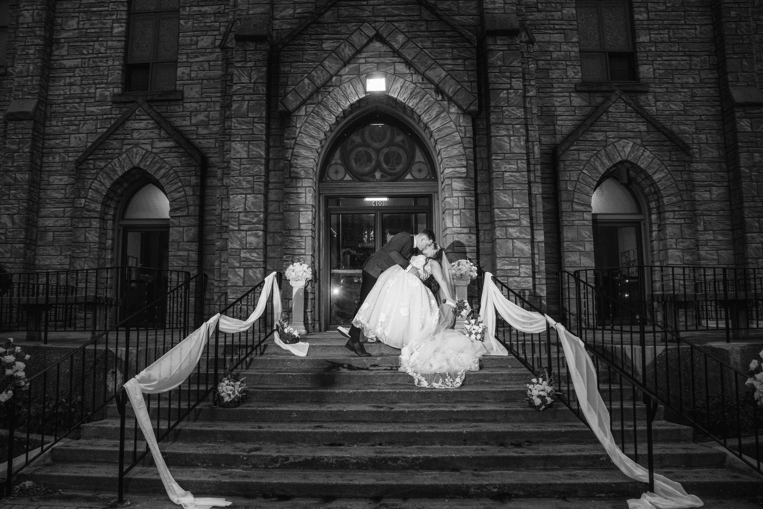 A couple in wedding attire kisses on decorated steps outside a church, at night—captured by a northwest Indiana and Chicago wedding photographer.