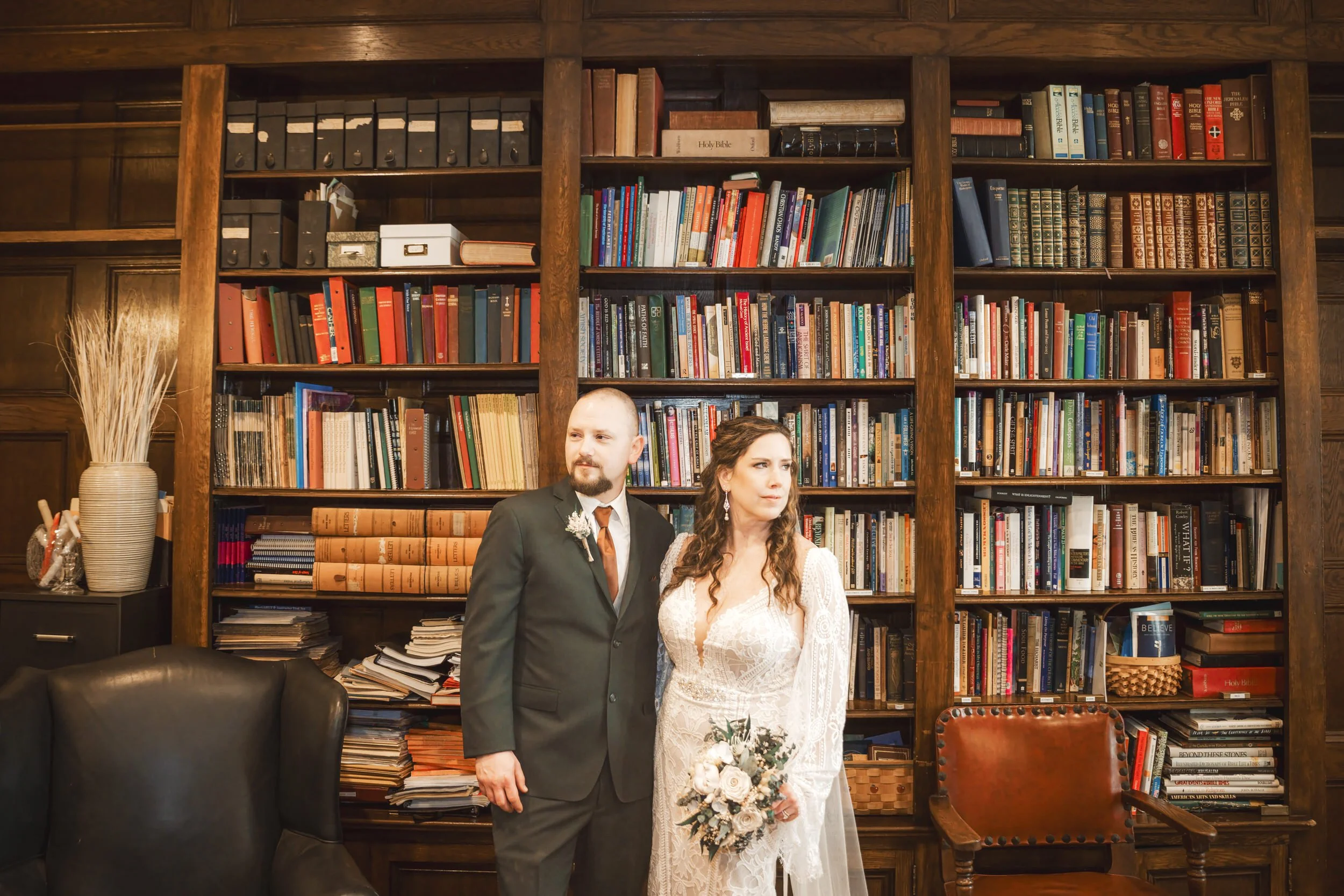 A bride and groom stand in front of an old world book case at Barker Hall at Trinity Church.