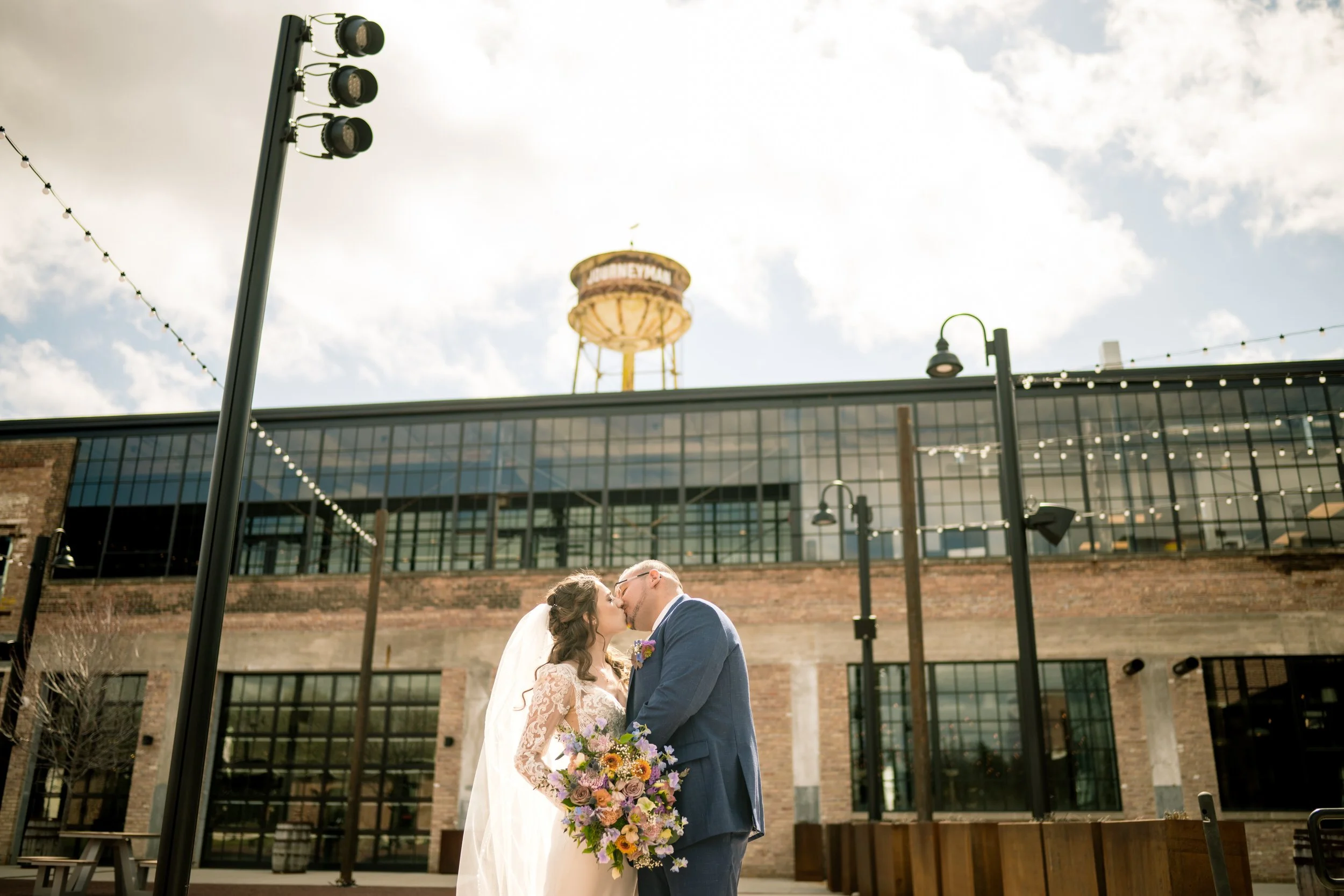 A bride and groom kiss outdoors in front of a brick industrial building with a water tower and string lights, captured by a northwest Indiana and Chicago wedding photographer.