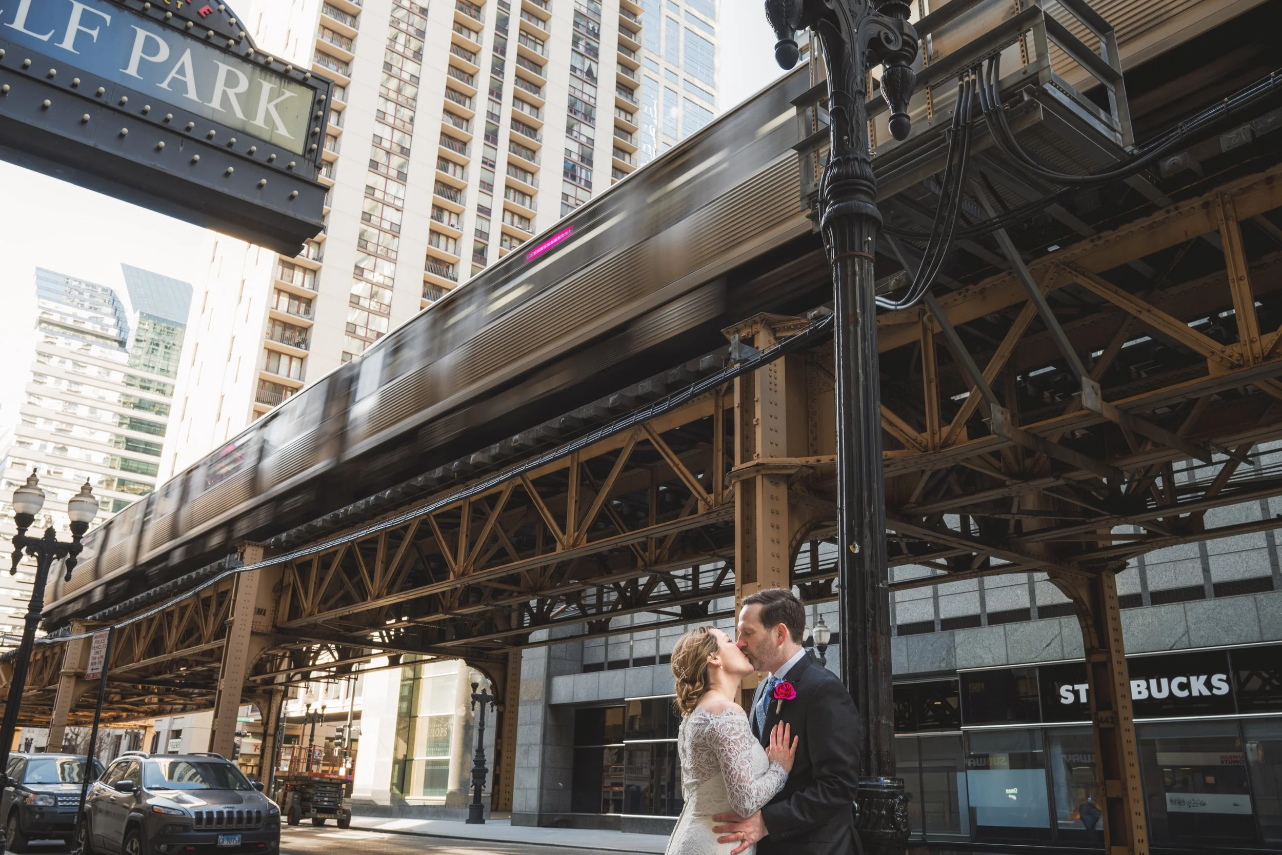 A bride and groom kiss on a city street beneath an elevated train track, with a moving train above and tall buildings in the background, captured by a Northwest Indiana and Chicago wedding photographer.