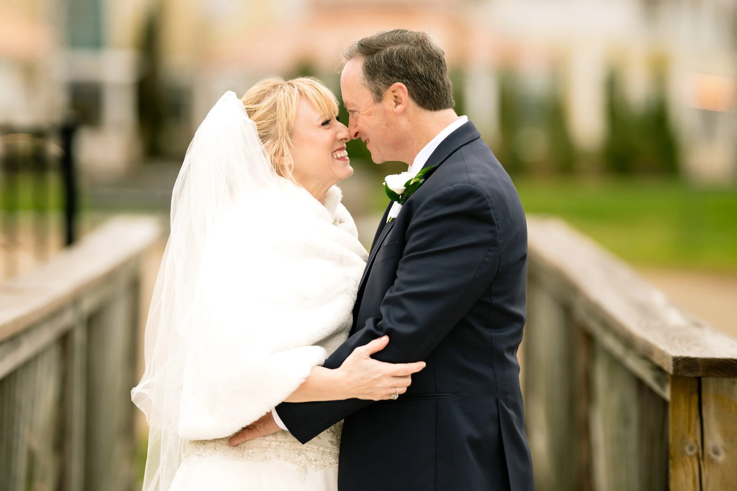A bride and groom stand close together on a wooden bridge, smiling at each other in formal wedding attire—captured by a talented northwest Indiana and Chicago wedding photographer.