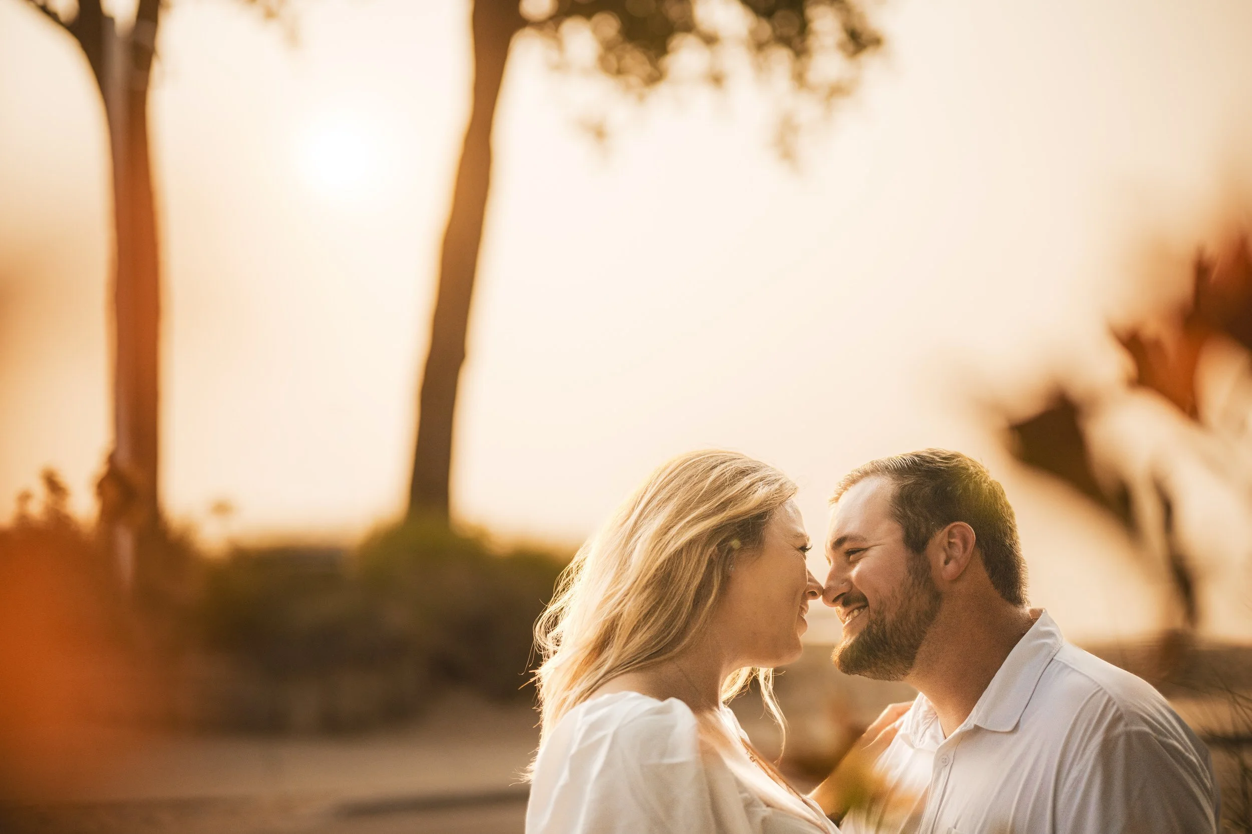 A couple smiling and touching foreheads outdoors during golden hour, with trees and soft sunlight in the background, beautifully captured by a southwest Michigan wedding photographer.