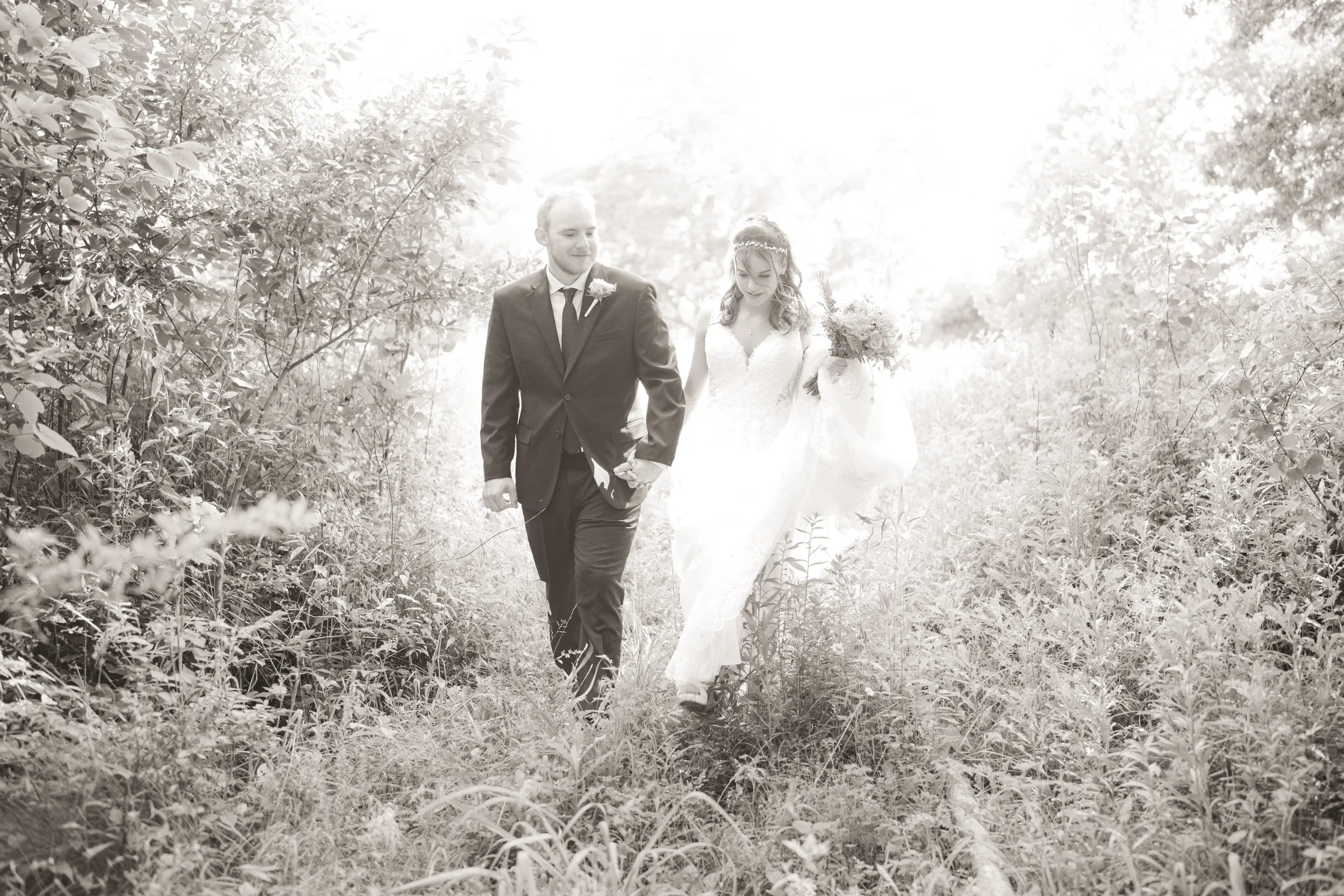 A bride and groom walk together through tall grass and foliage, dressed in wedding attire under bright natural light—captured by a Northwest Indiana and Chicago wedding photographer.