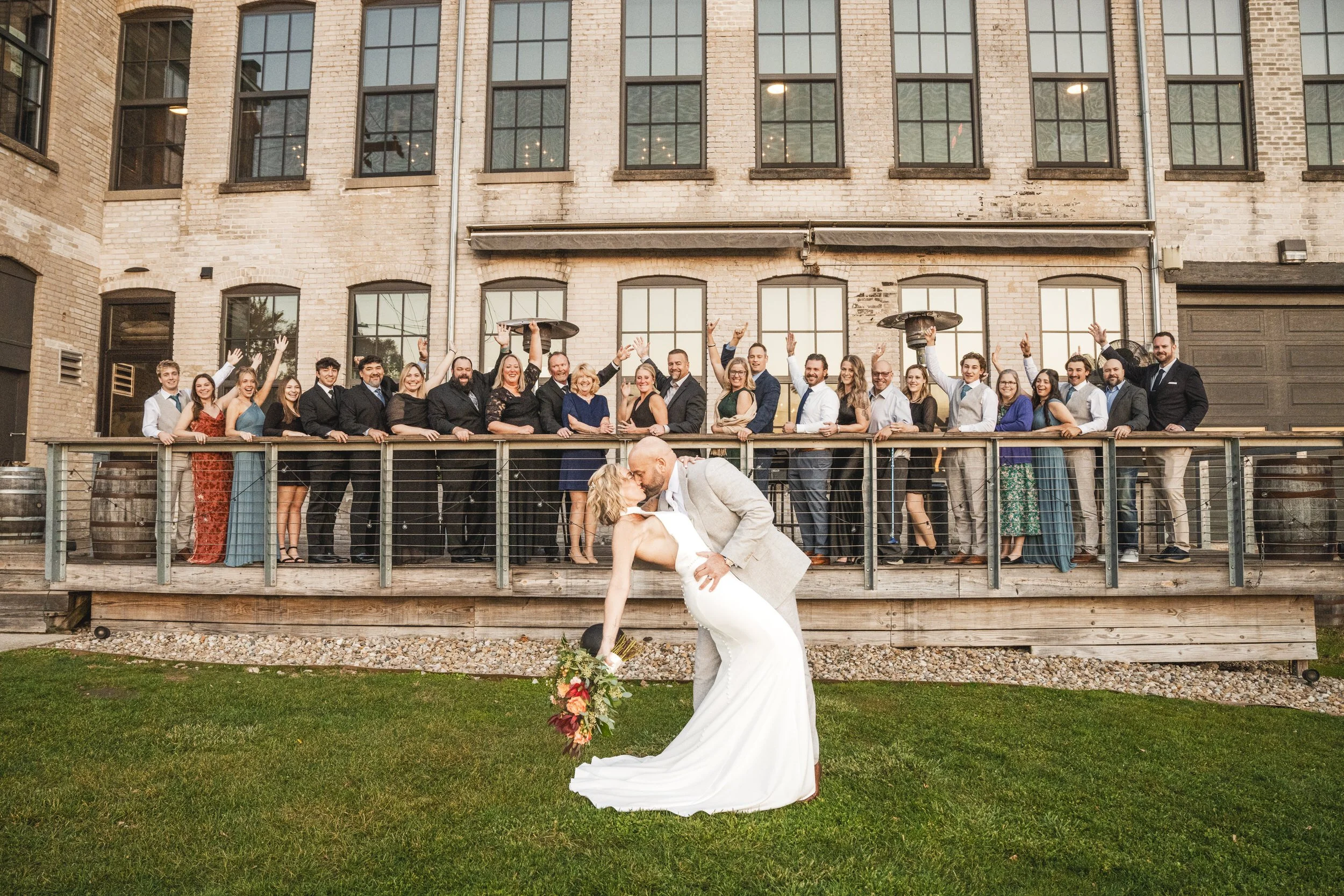 A wedding groups photo with the bride and groom out front dipping as family and friends cheer from behind them.