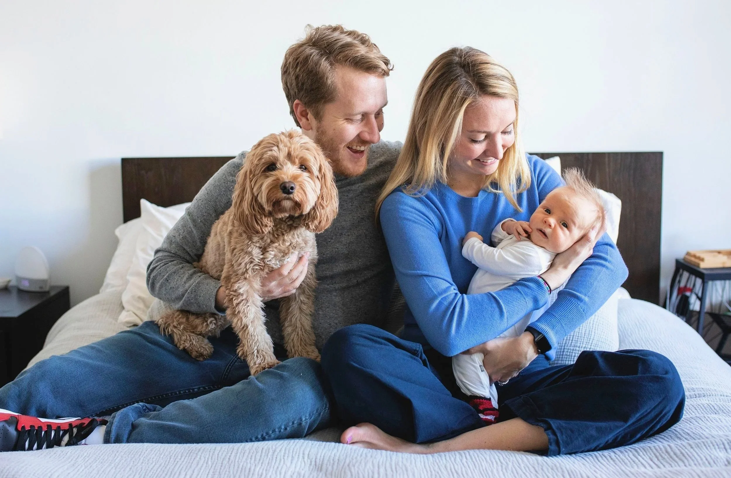 A family sitting on a bed with a dog, a woman holding a baby, and a man holding the dog, all smiling and looking happy. Photographed by family photographer Marisa S. Cohen.