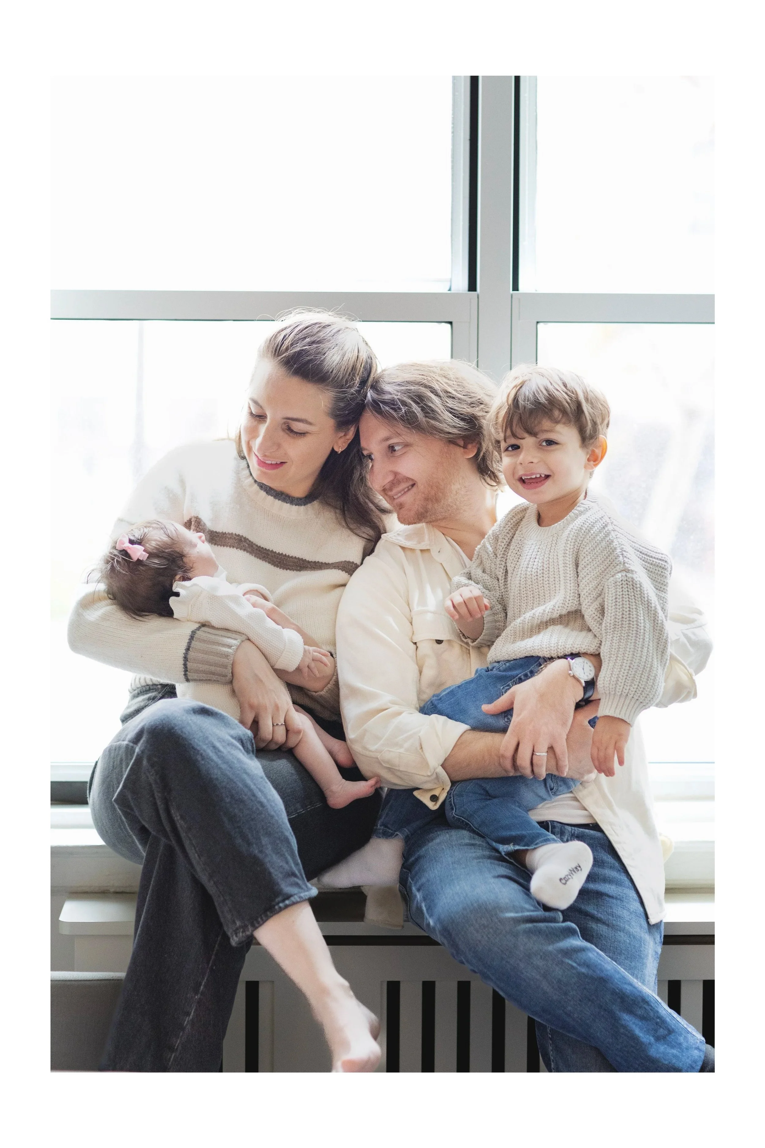 Family of five sitting by a large window, holding and smiling at their newborn baby and young boy. Photographed by NYC family photographer Marisa S. Cohen.