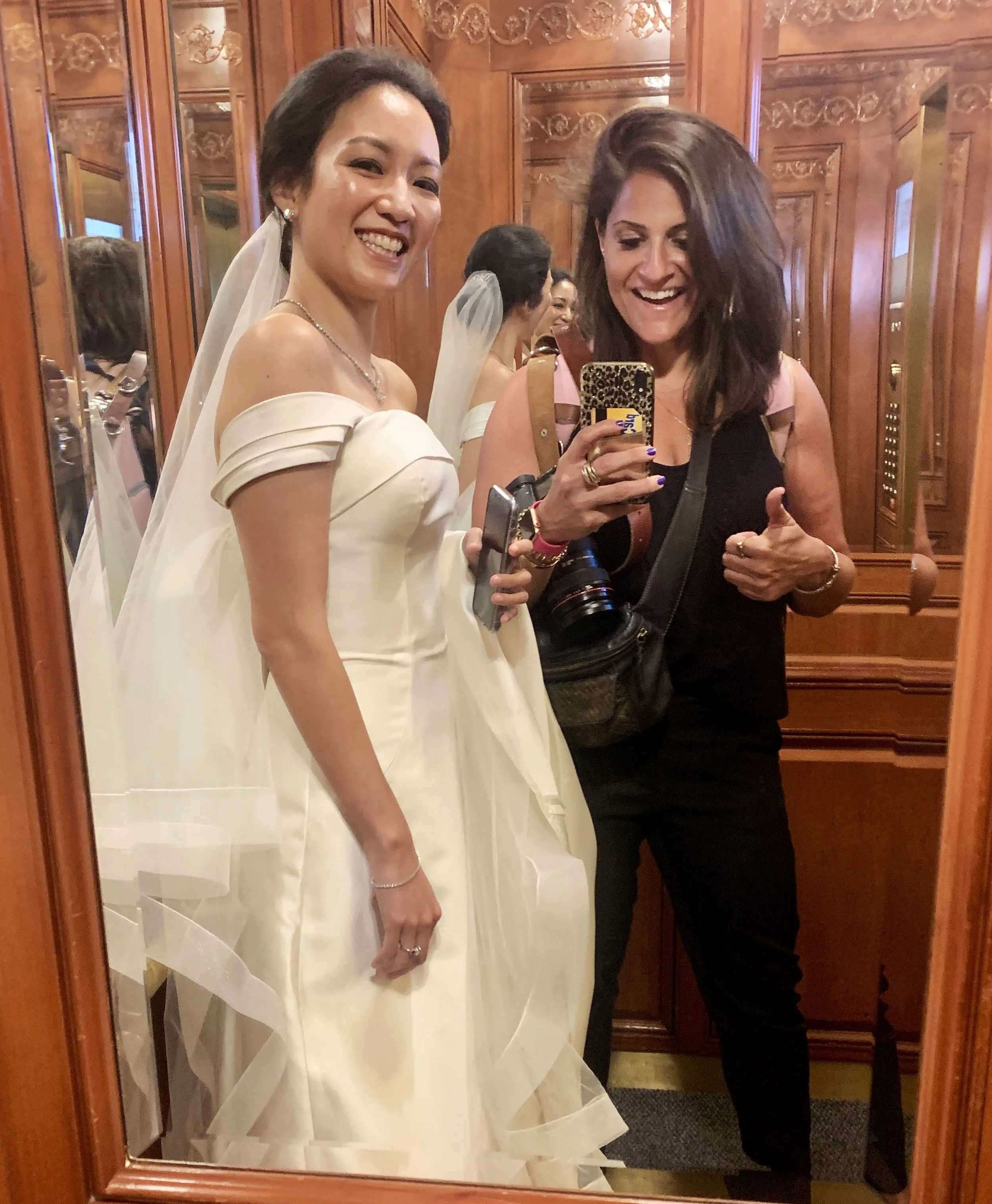 A woman in a wedding dress taking a mirror selfie with another woman in black attire inside an ornate wooden elevator, both smiling. Photographed by family photographer Marisa S. Cohen.