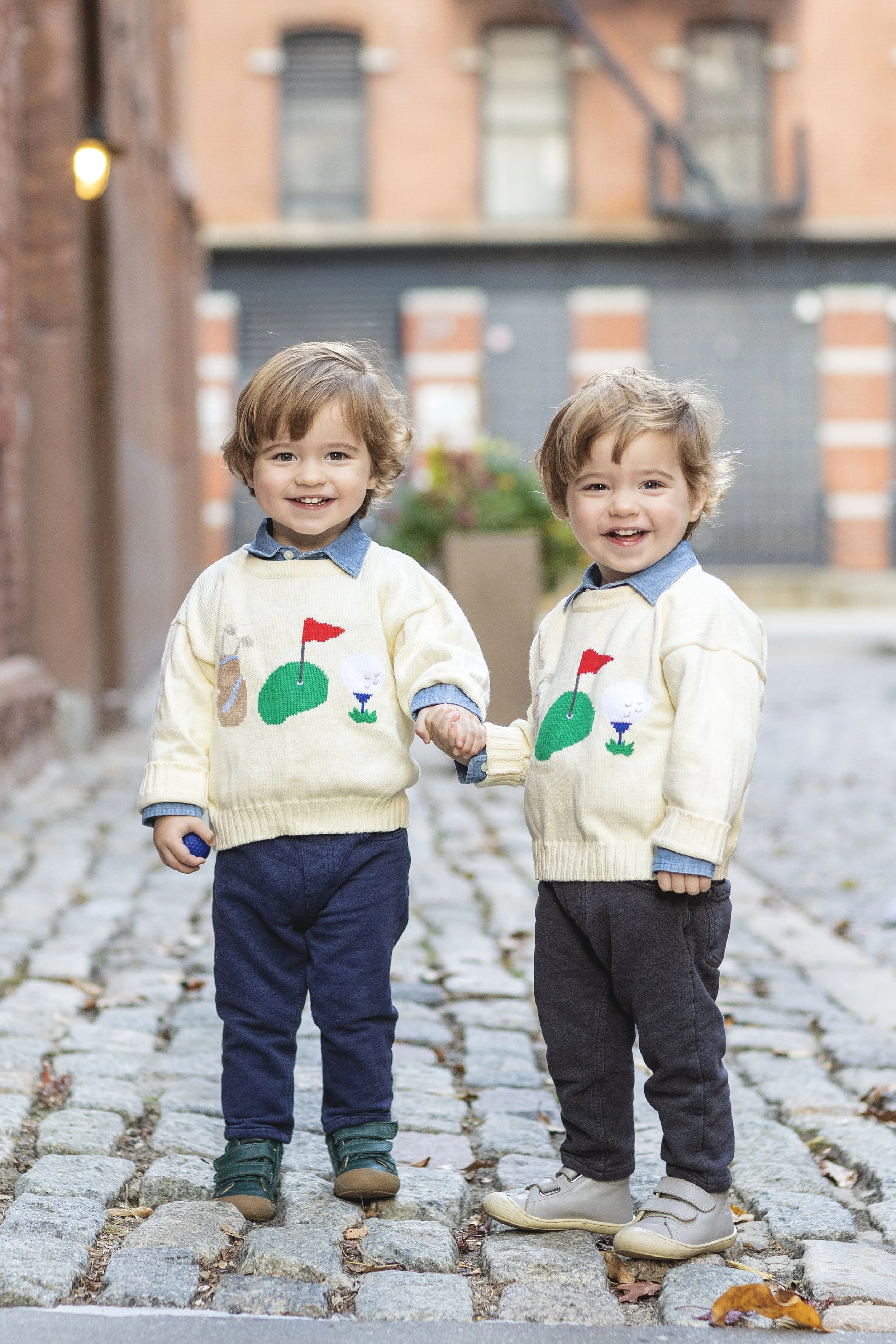 Two young boys holding hands, standing on a cobblestone street, wearing matching cream sweaters with golf course designs and blue collared shirts underneath. Photographed by family photographer Marisa S. Cohen.
