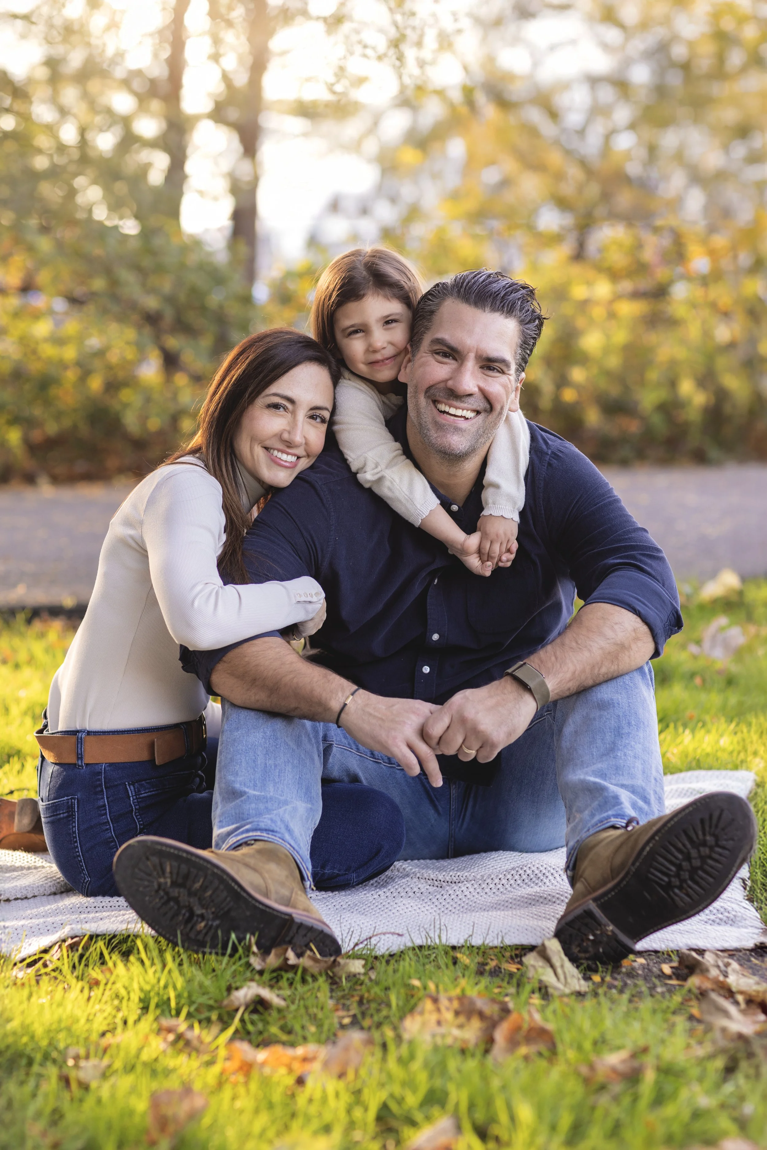 A happy family of three sitting on a blanket outdoors during autumn, with trees and fall foliage in the background. Photographed by family photographer Marisa S. Cohen.