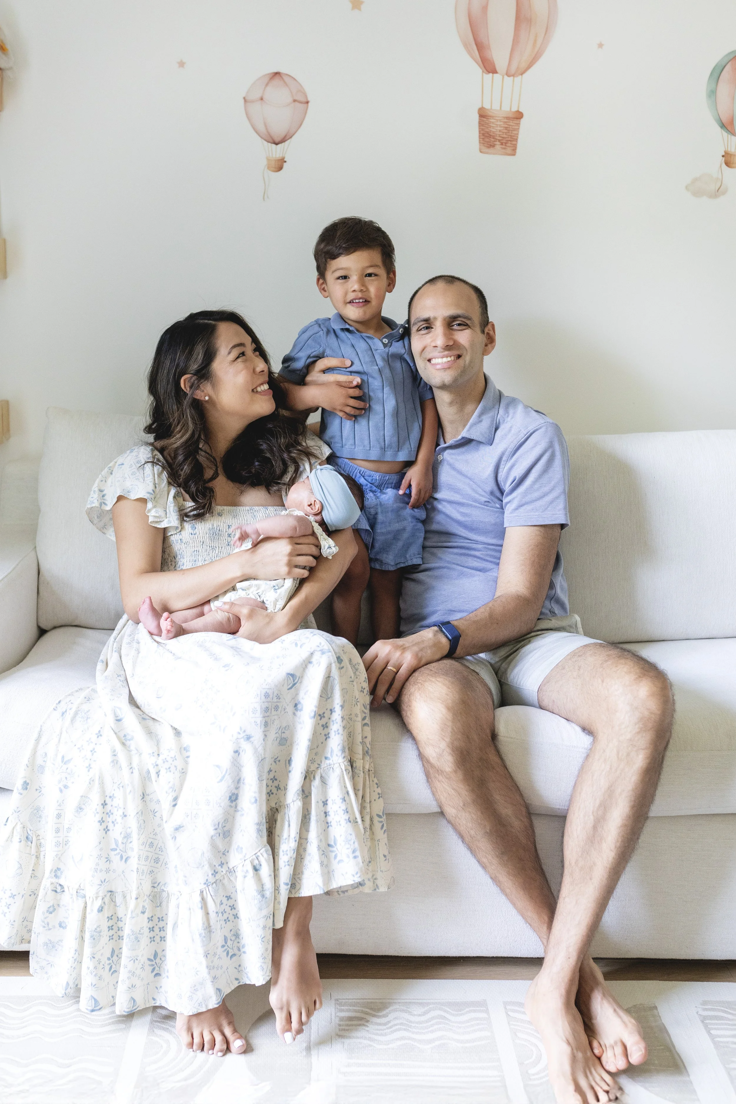 Family of four sitting on a white couch, with a woman holding a newborn baby, a young boy standing on the couch, and a man sitting with one leg crossed over the other. Photographed by family photographer Marisa S. Cohen.