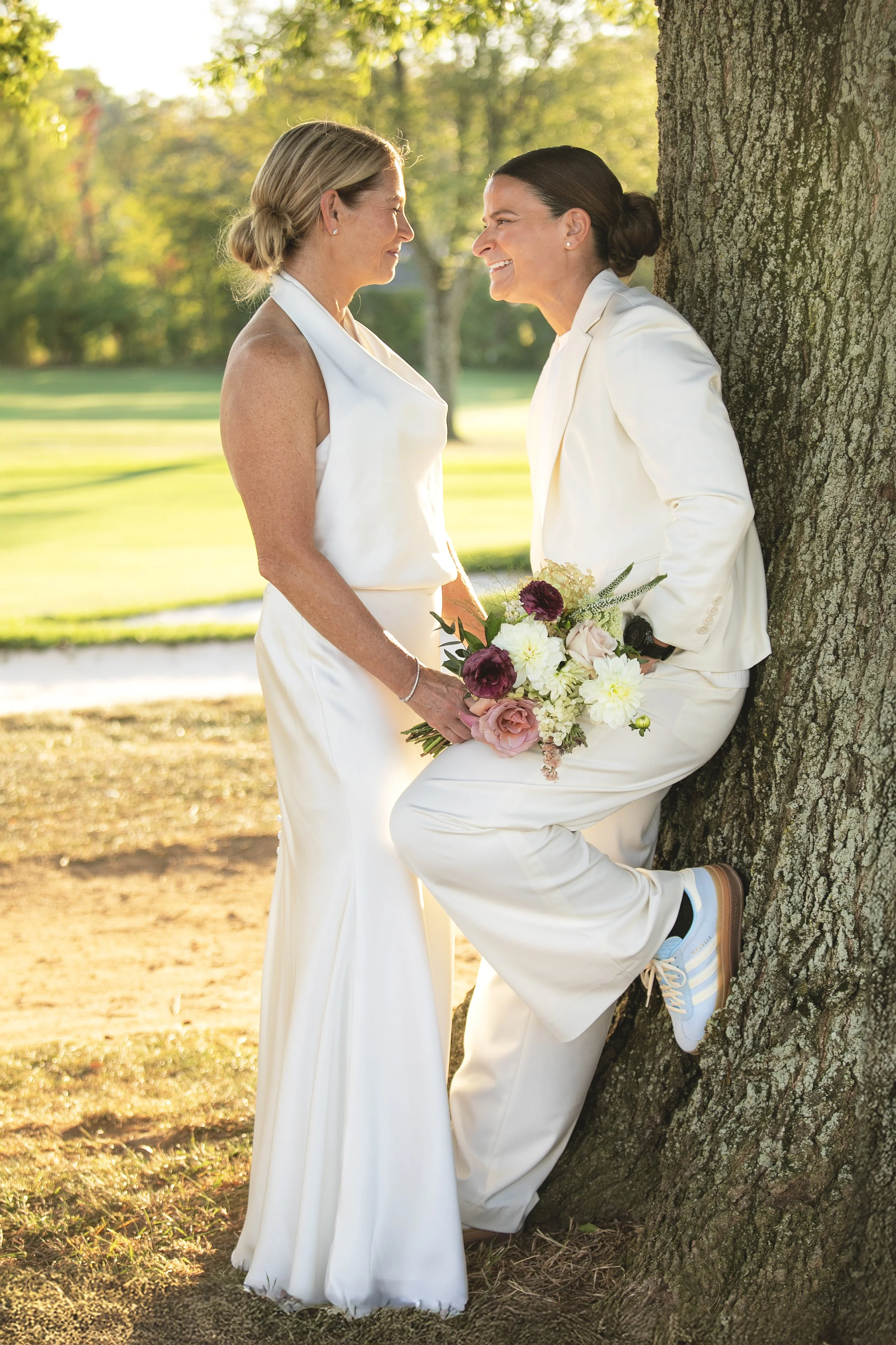 Two women, one with short blonde hair and the other with dark hair in a bun, dressed in white, smiling and looking at each other while holding a bouquet of flowers outdoors near a tree, with green trees and grass in the background.
