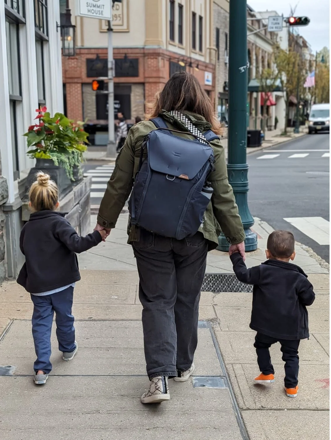 A woman walking down the street holding hands with two young children, one on each side. The woman has a large backpack and is wearing a green jacket and patterned boots. The children are dressed in dark jackets, with one child wearing orange shoes and the other sneaking a peek at the camera. The scene takes place on a city sidewalk with buildings, flowers, and traffic in the background.