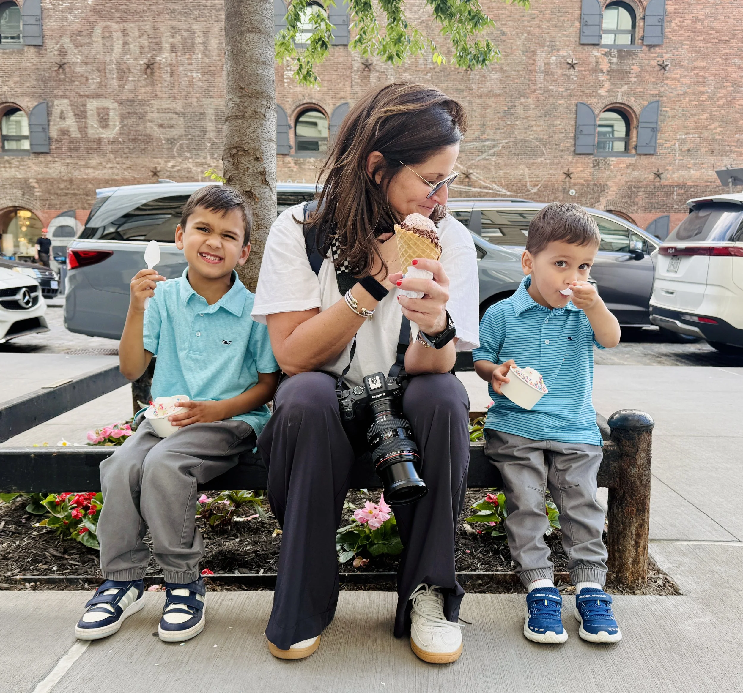 A woman with three young children sitting on a bench enjoying ice cream and cups of frozen yogurt outdoors in an urban setting with parked cars and brick buildings in the background. Photographed by family photographer Marisa S. Cohen.