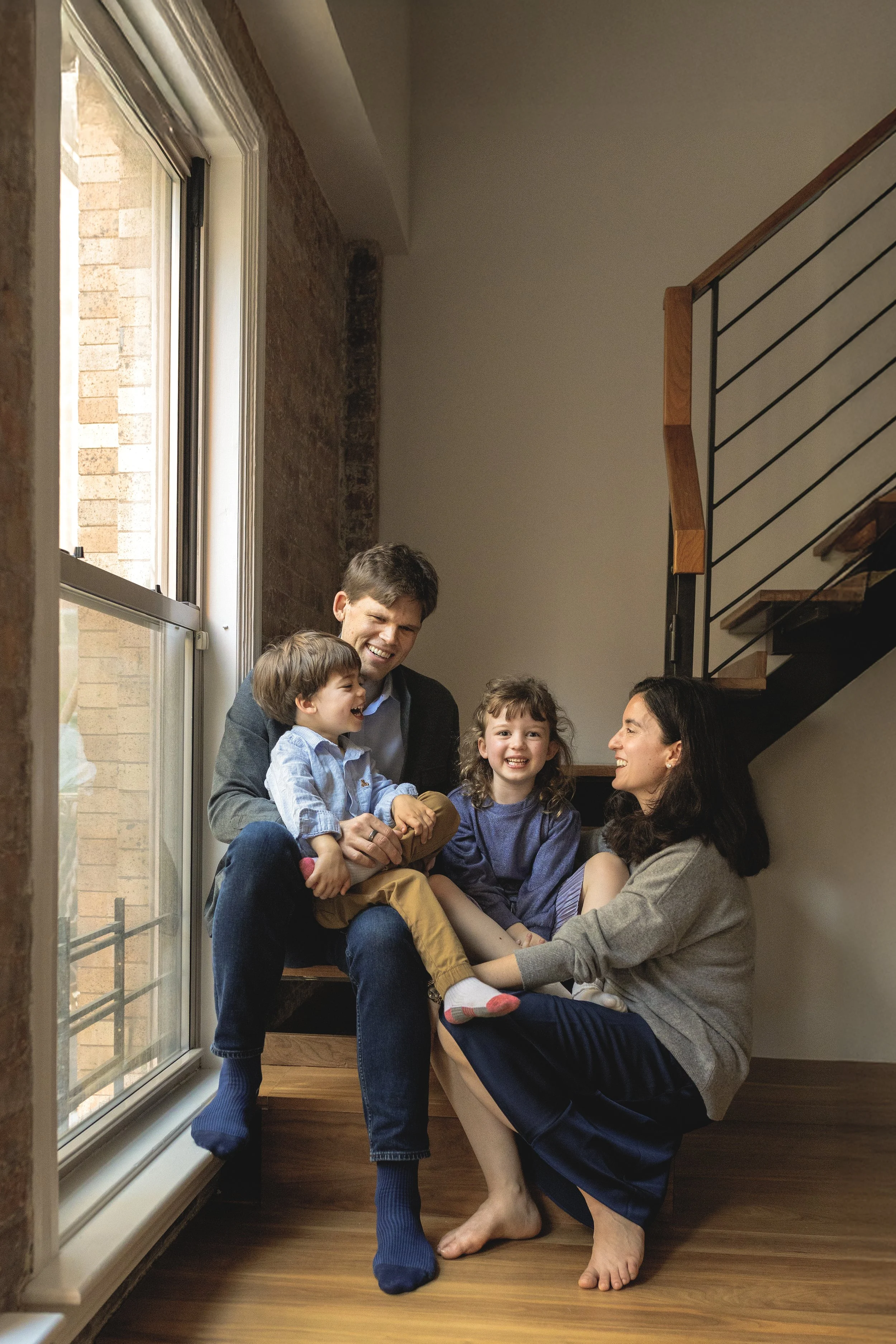 A family of five sits on a staircase by a large window, smiling and laughing together indoors. Photographed by family photographer Marisa S. Cohen.
