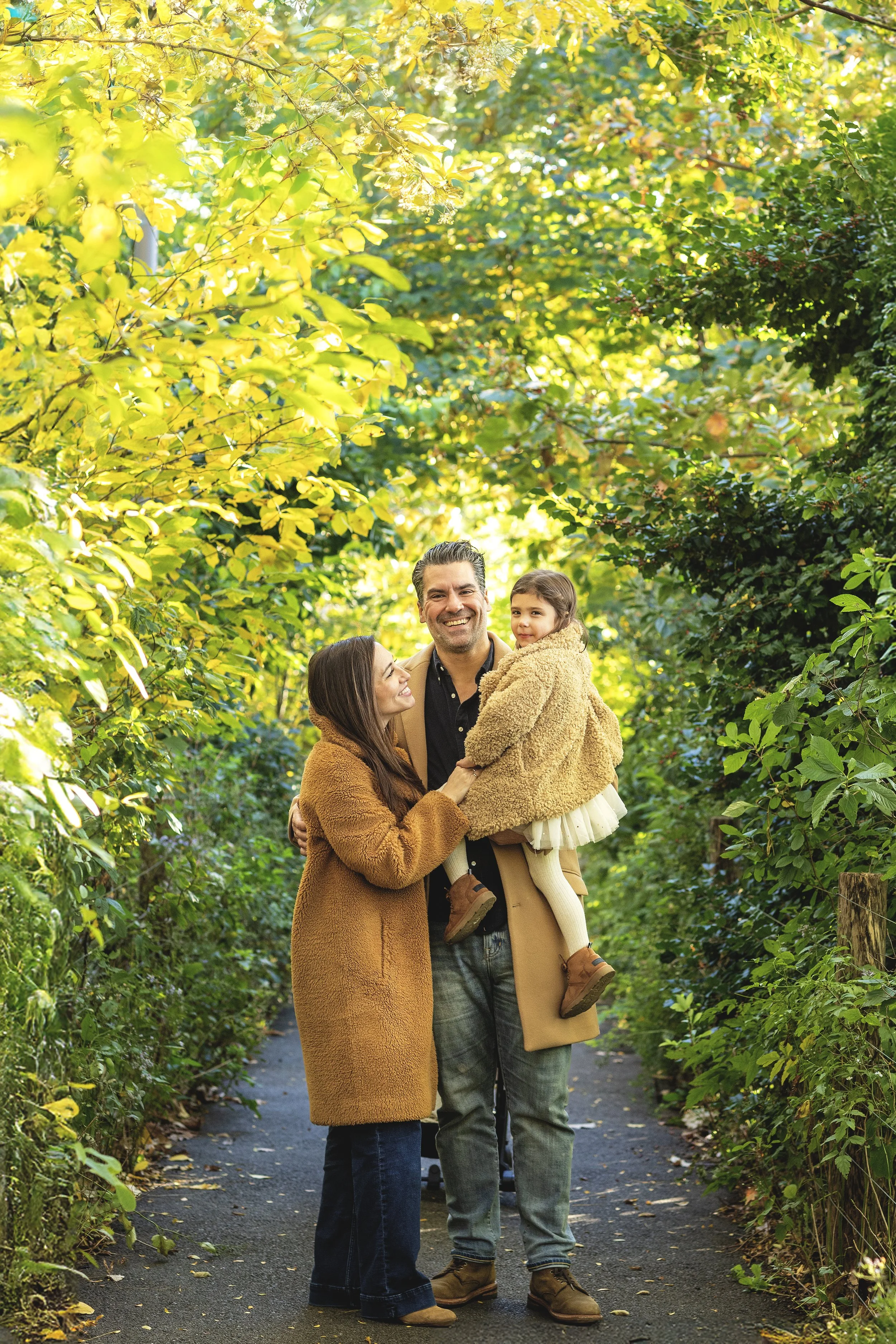 A family of three, including a man, a woman, and a young girl, standing on a path surrounded by lush green trees. They are smiling and embracing each other in a cheerful outdoor setting.