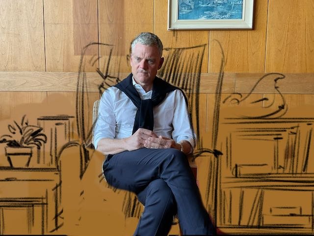 And now, here’s Patrick as the Therapist sitting in the correct high-back chair, in front of a low bookcase. And there’s art on the wall. And next to the Therapist is a small table with a small plant on it.