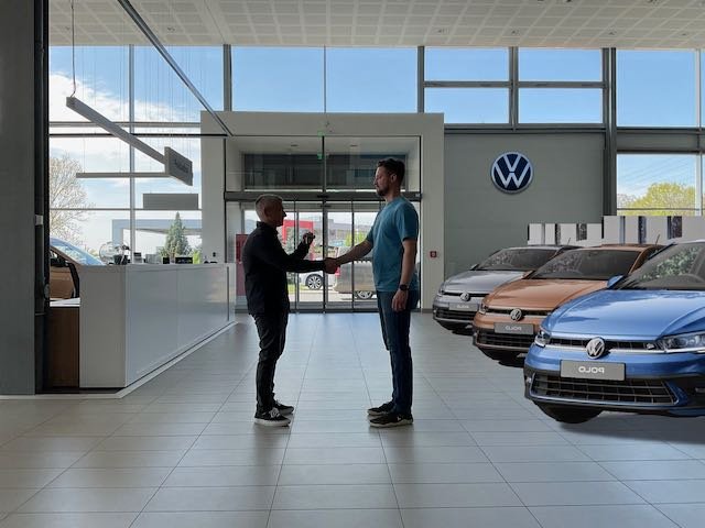 …Michael, as 33-year-old Maxime, shaking hands with the dealer while a large VW LOGO sits in the background, along with cars parked at an angle. The cars could also be parked without an angle. It’s whatever everyone likes and thinks looks best.