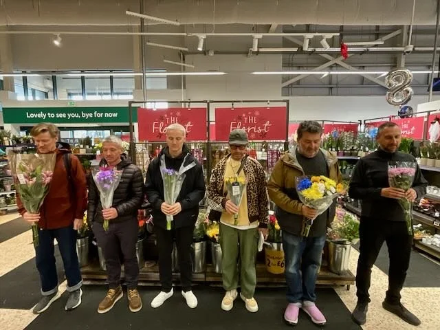 …to a WIDE of the FLOWERS SECTION, where the FIVE YOUNG LADS are standing in a line, FACING camera. Each is STARING DOWN at their own UNIQUE BOUQUET, as the MORRISONS FLORIST looks on. In perfect SYNCHRONICITY… 